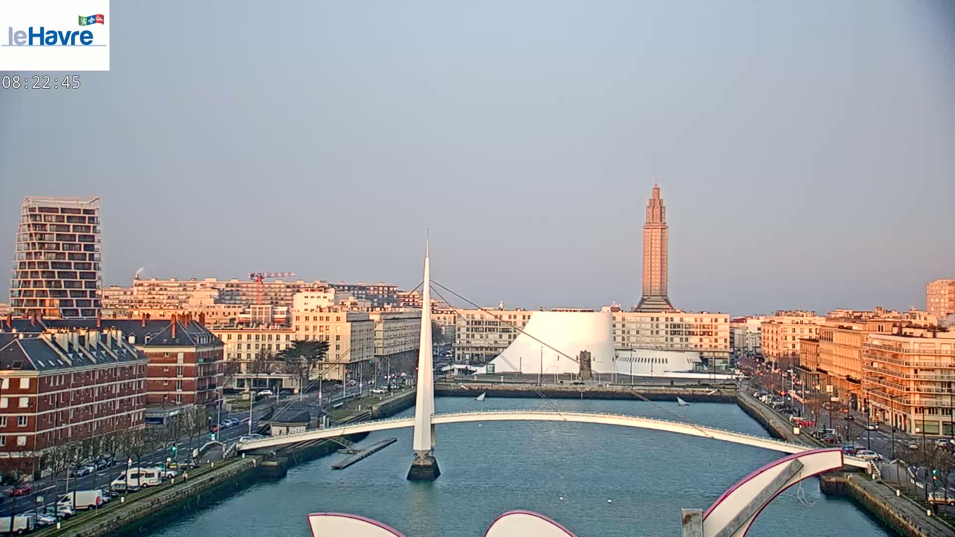 A cityscape with a modern bridge spanning a waterway, under a clear, pale sky, features a tall tower and various buildings along the waterfront.