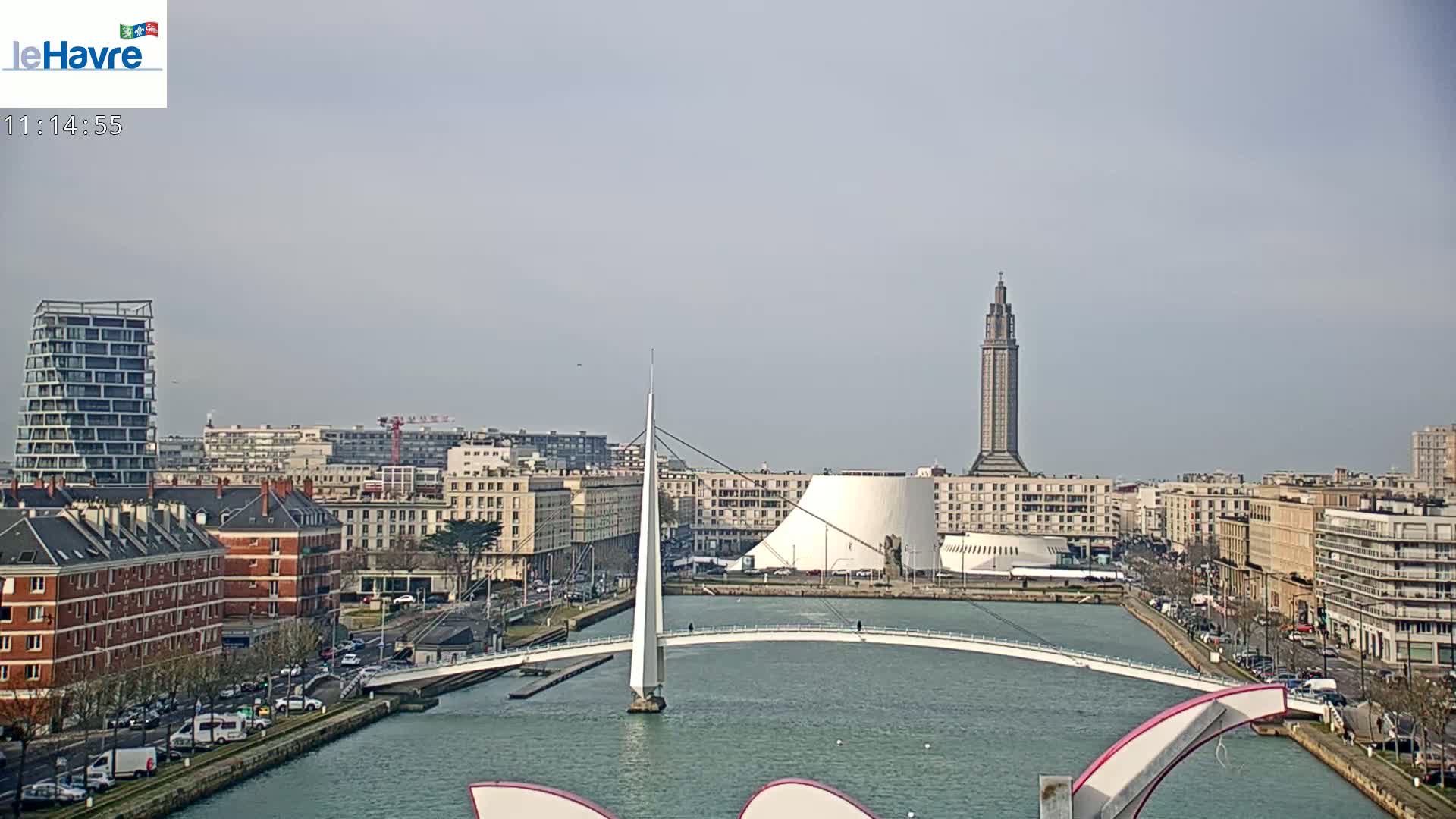 A cityscape with a modern white bridge over a waterway, under a mostly clear sky, features a tall tower and other buildings.