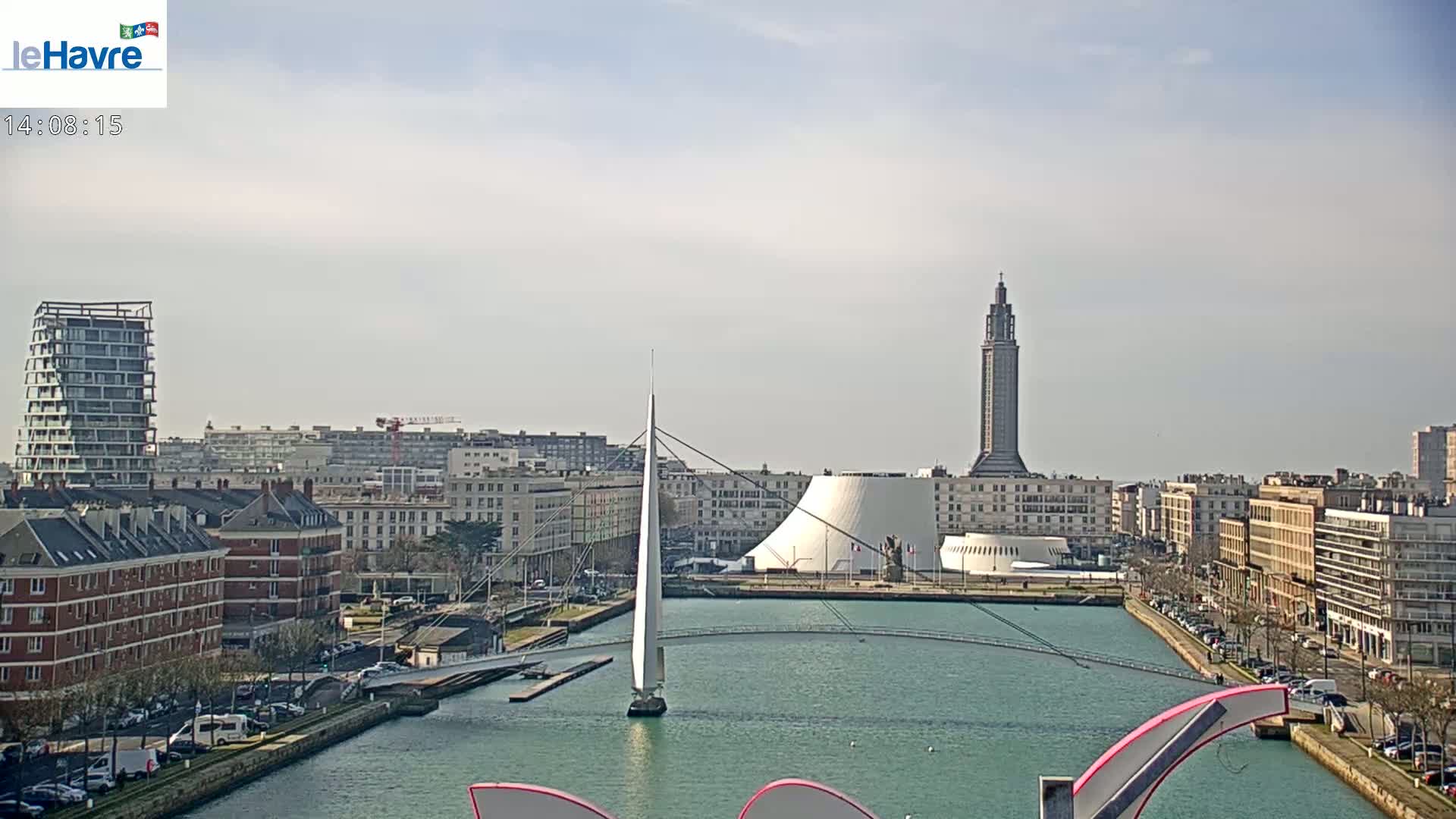 A sailboat is in a waterway next to a city with several buildings, including a tall tower and a domed structure, under a mostly clear sky.
