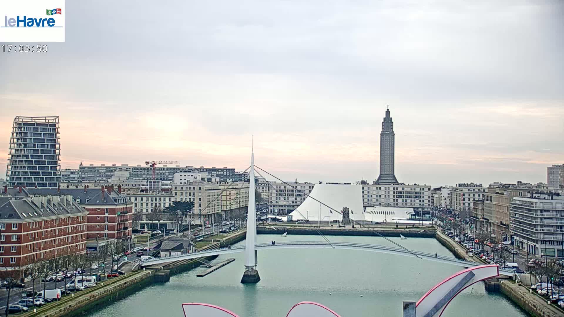 A cityscape with a modern bridge over a calm waterway, under a pale, overcast sky at sunset.