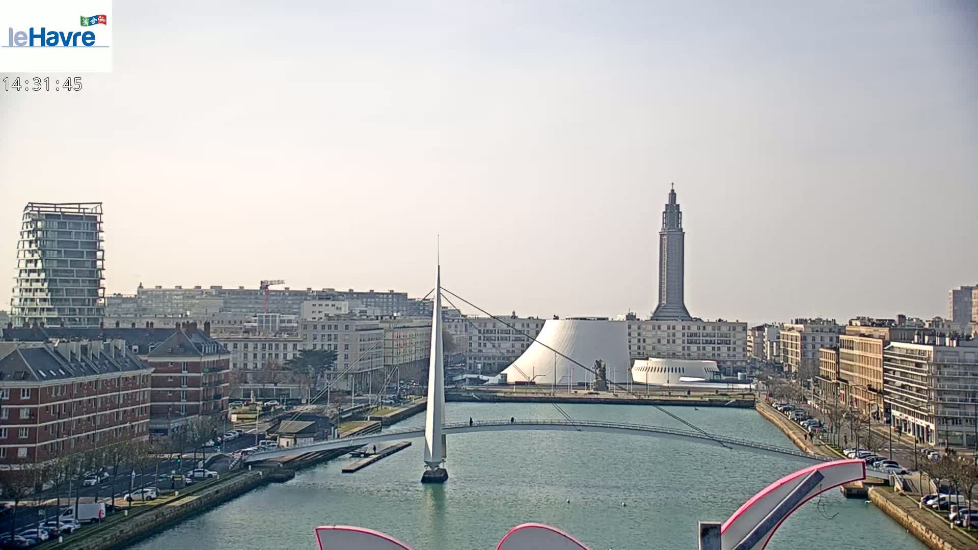 A sunny cityscape shows a canal with a sailboat under a white pedestrian bridge,  modern buildings, and a tall tower in the background.