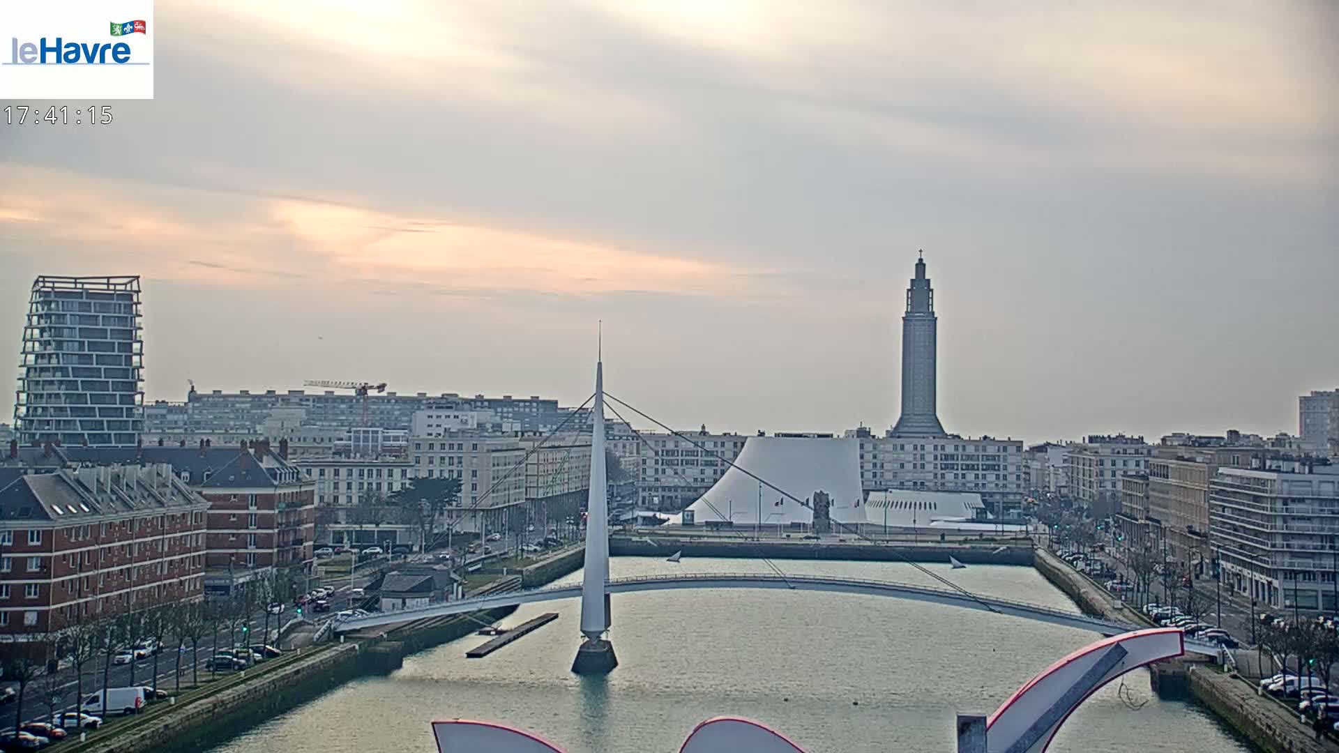 A city waterfront at sunset shows a tall tower, a modern white building, a slender bridge, and various buildings lining a canal.