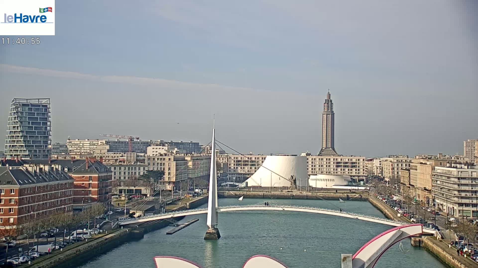 A city waterfront on a sunny day features a white pedestrian bridge, modern buildings, and a tall tower in the distance.