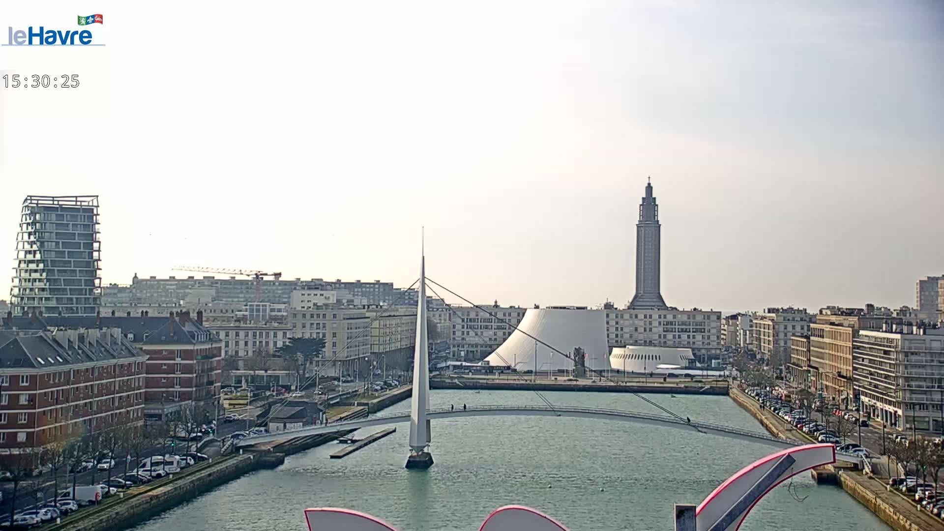A city waterfront on a partly sunny day features a white pedestrian bridge crossing a canal, with buildings and a tall tower in the background.