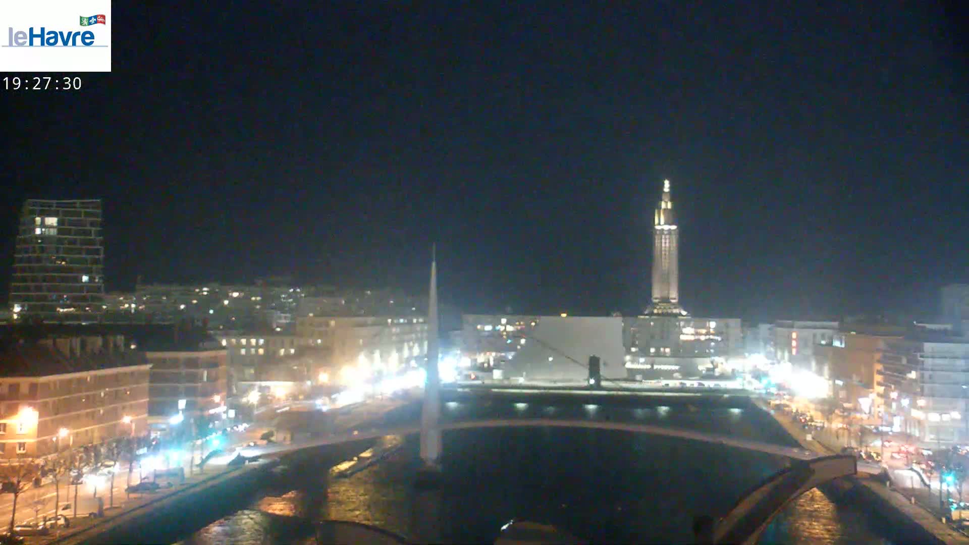 A nighttime cityscape with a tall, illuminated tower in the center, a canal, and a bridge, under a clear, dark sky.