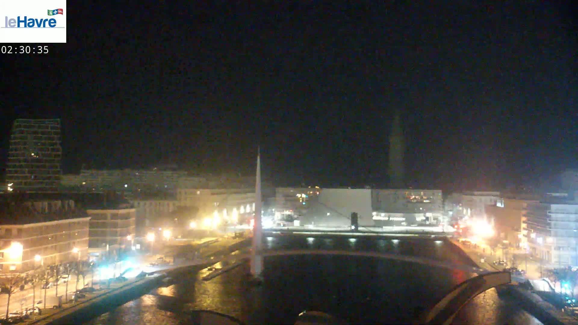 A nighttime view of a city waterfront with buildings, streetlights, and a tall, slender structure illuminated against a dark sky.