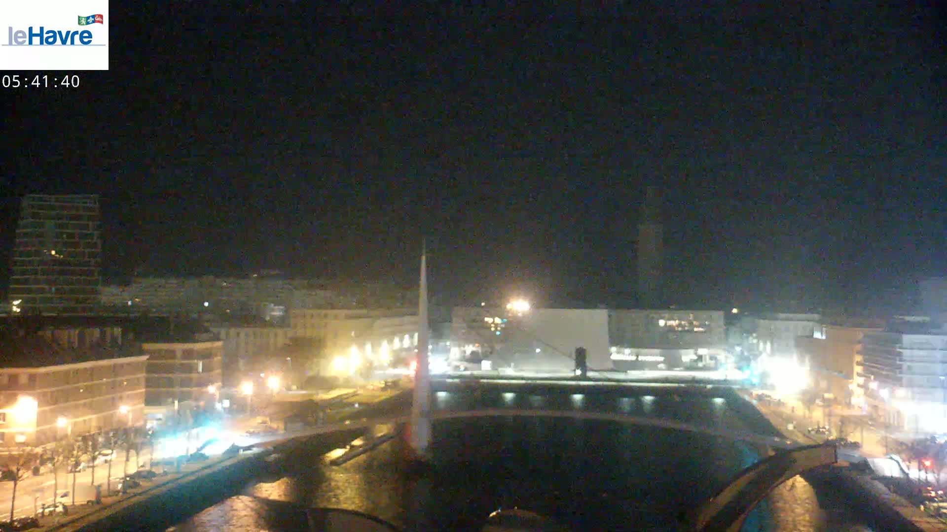 A nighttime view of a city waterfront with a bridge and buildings illuminated by streetlights under a dark sky.