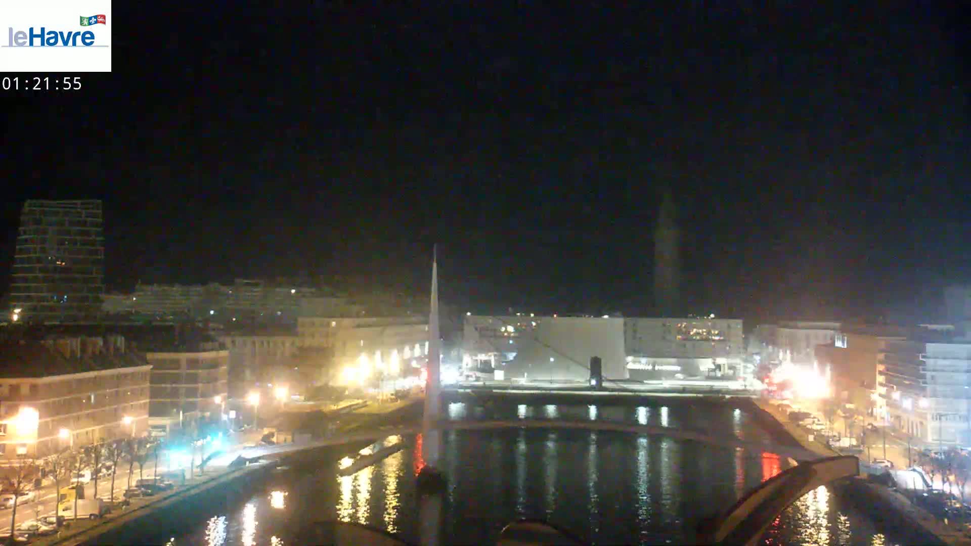 A nighttime view of a city waterfront, showing buildings lit up along a canal with a tall structure in the background under a clear, dark sky.