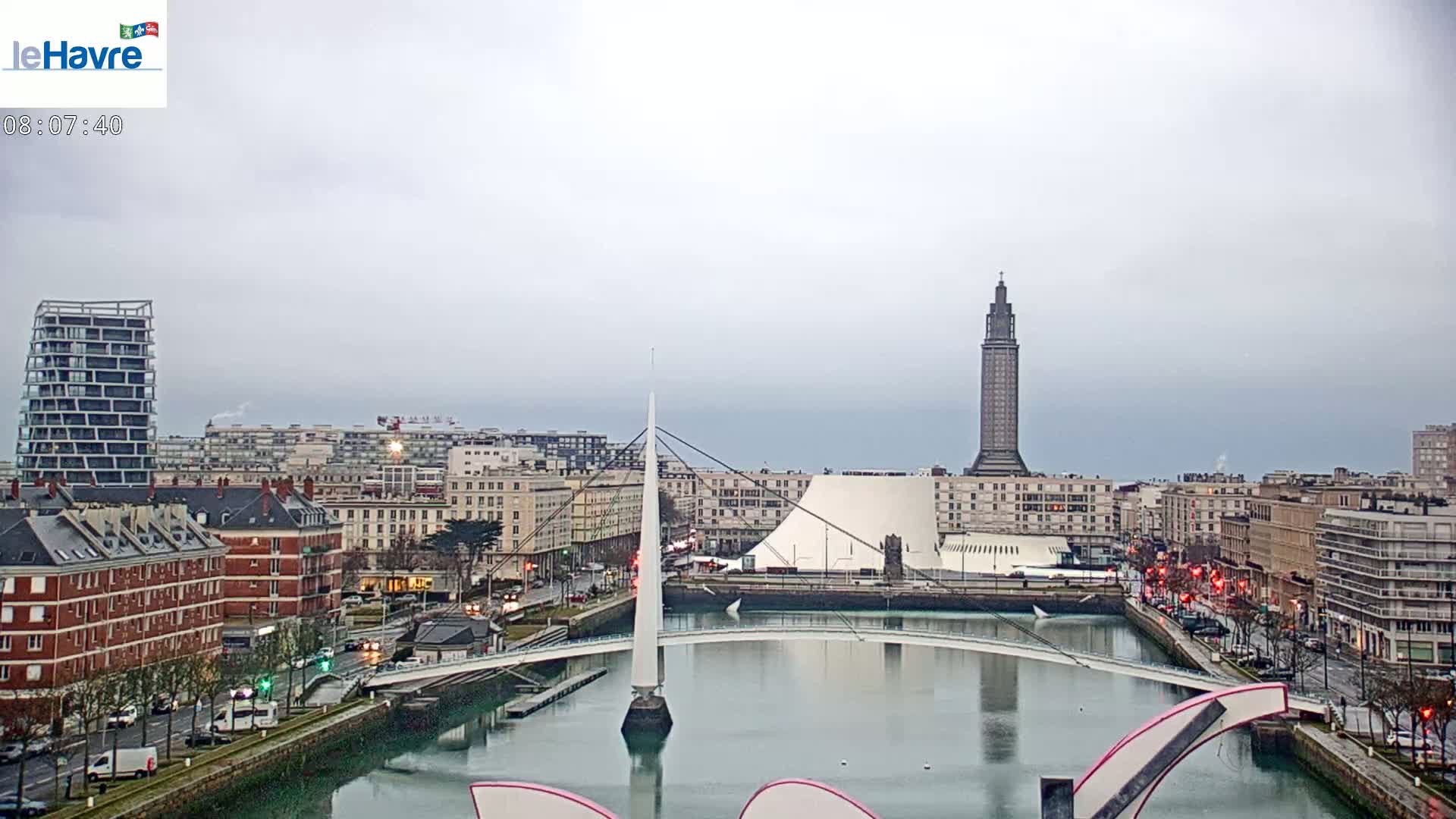 A city harbor scene on an overcast day shows a white pedestrian bridge spanning a calm waterway, with modern and older buildings lining the waterfront, and a tall tower in the distance.