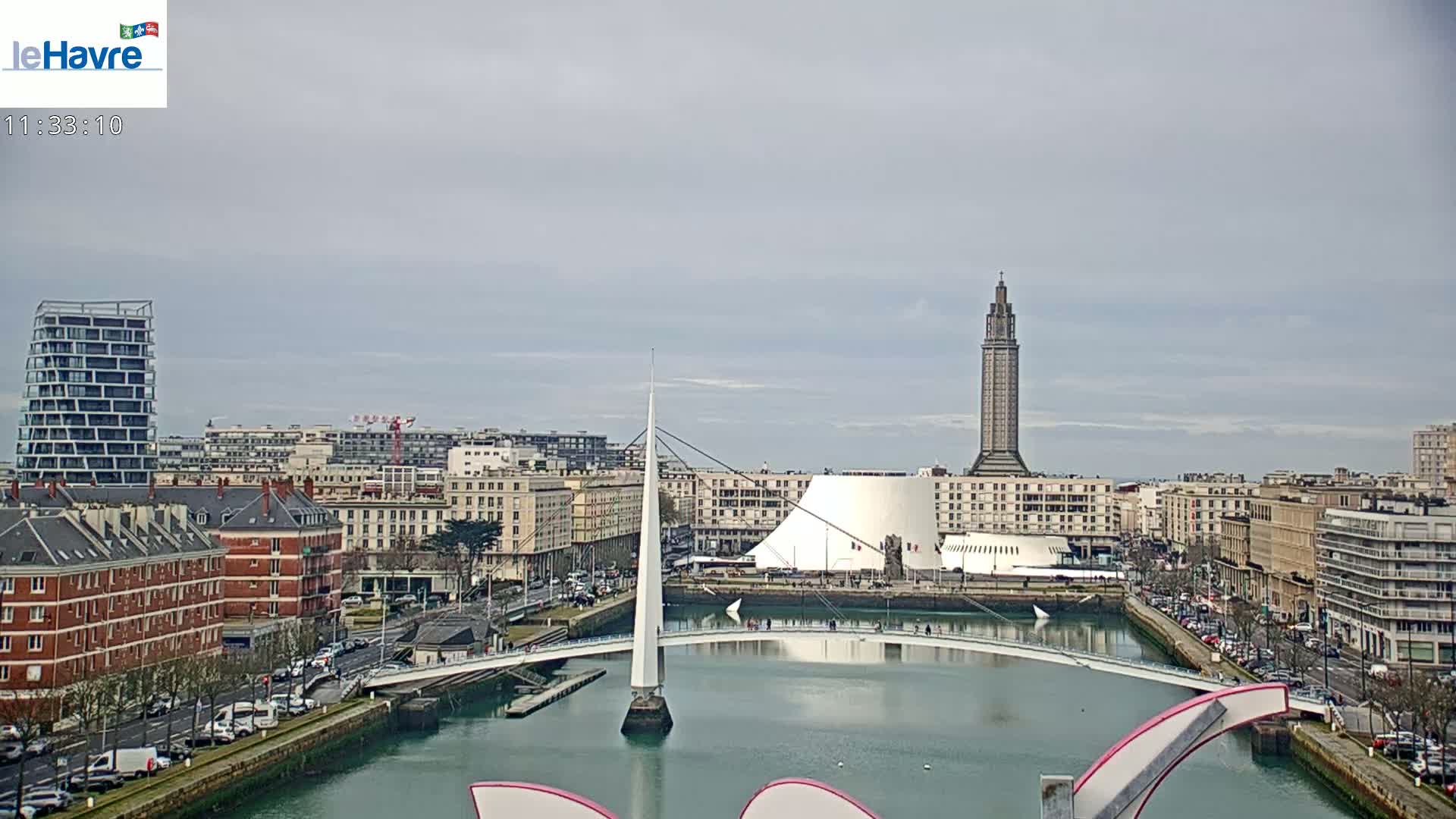A city waterfront scene under an overcast sky shows a modern white bridge spanning a waterway, with various buildings, including a tall tower, in the background.