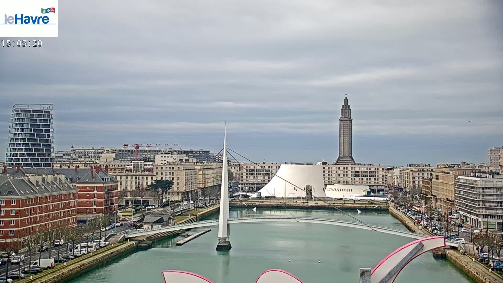 A city skyline with overcast skies shows a modern bridge spanning a waterway, leading to a tall tower and various buildings, including a large, white, curved structure.