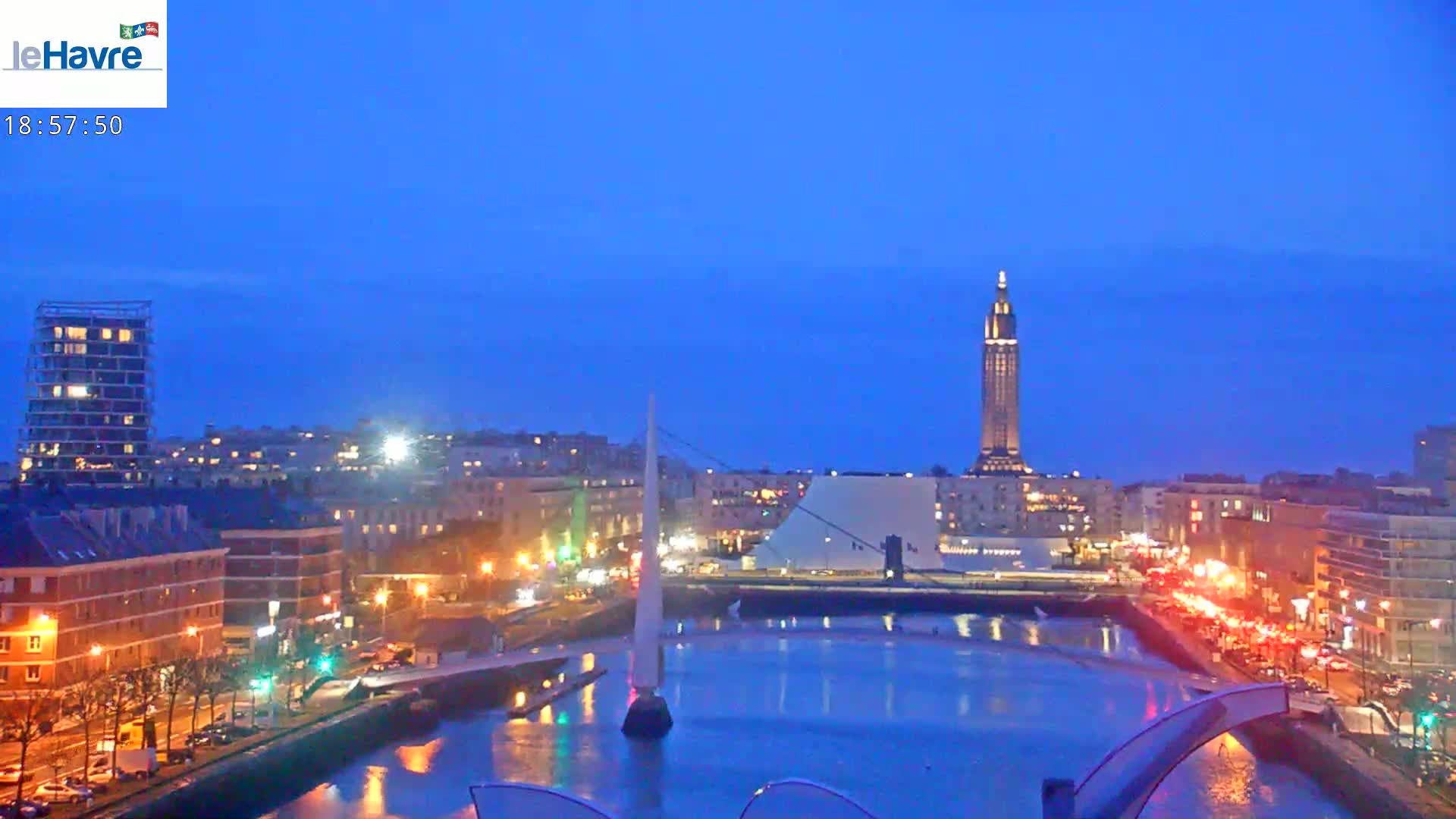 A city at twilight, with a calm body of water reflecting the lights of buildings and vehicles along the shore, under a clear, dark blue sky.