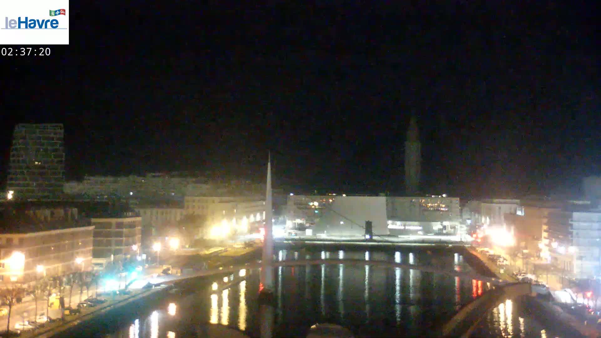 A nighttime view of a city waterfront, showing buildings illuminated along a calm waterway reflecting city lights under a dark sky.