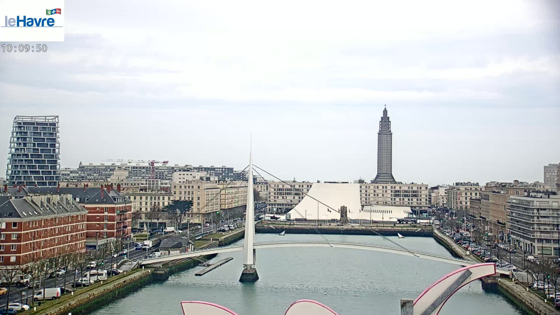 A city skyline with a tall tower and modern buildings is seen across a waterway, with a slender white bridge connecting the foreground to the opposite shore, under an overcast sky.