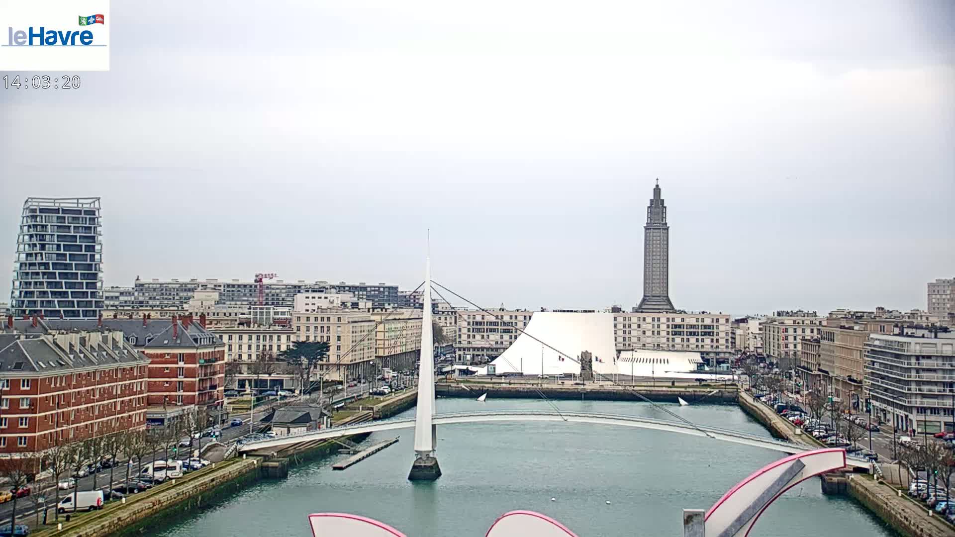 A cityscape under an overcast sky features a white cable-stayed bridge spanning a waterway, with a tall tower and modern buildings in the background.