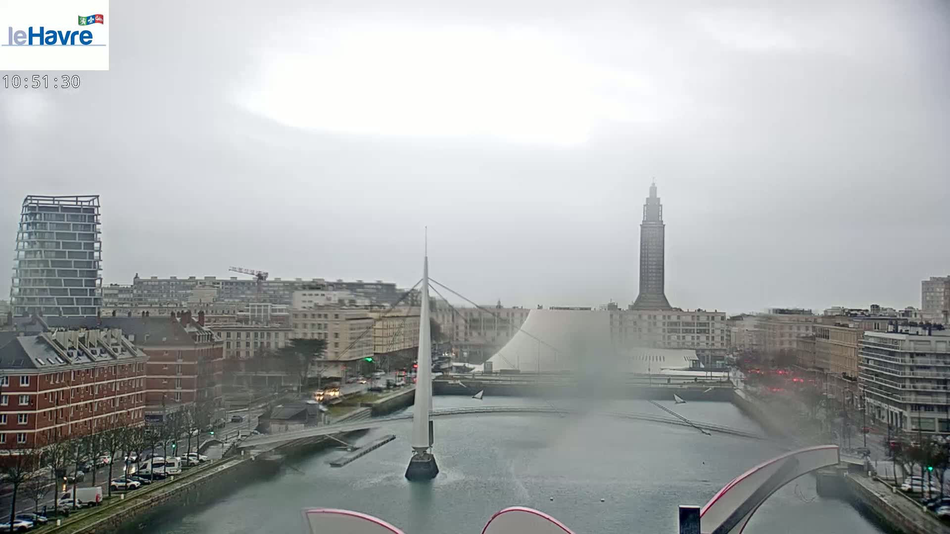 A city waterfront view on an overcast day shows a canal, a bridge, several buildings, and a tall tower in the distance.