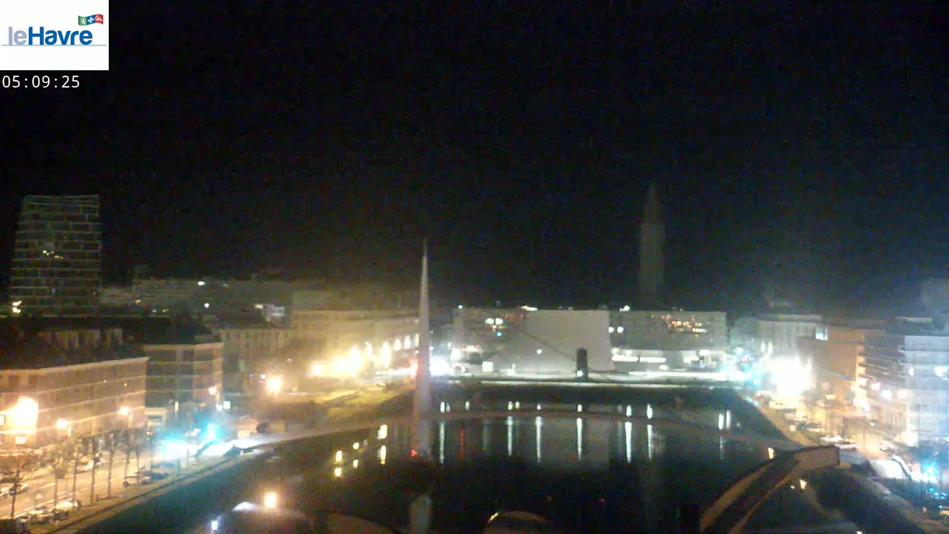 A nighttime city scene shows a body of water reflecting the lights of buildings along its banks, under a dark, clear sky.