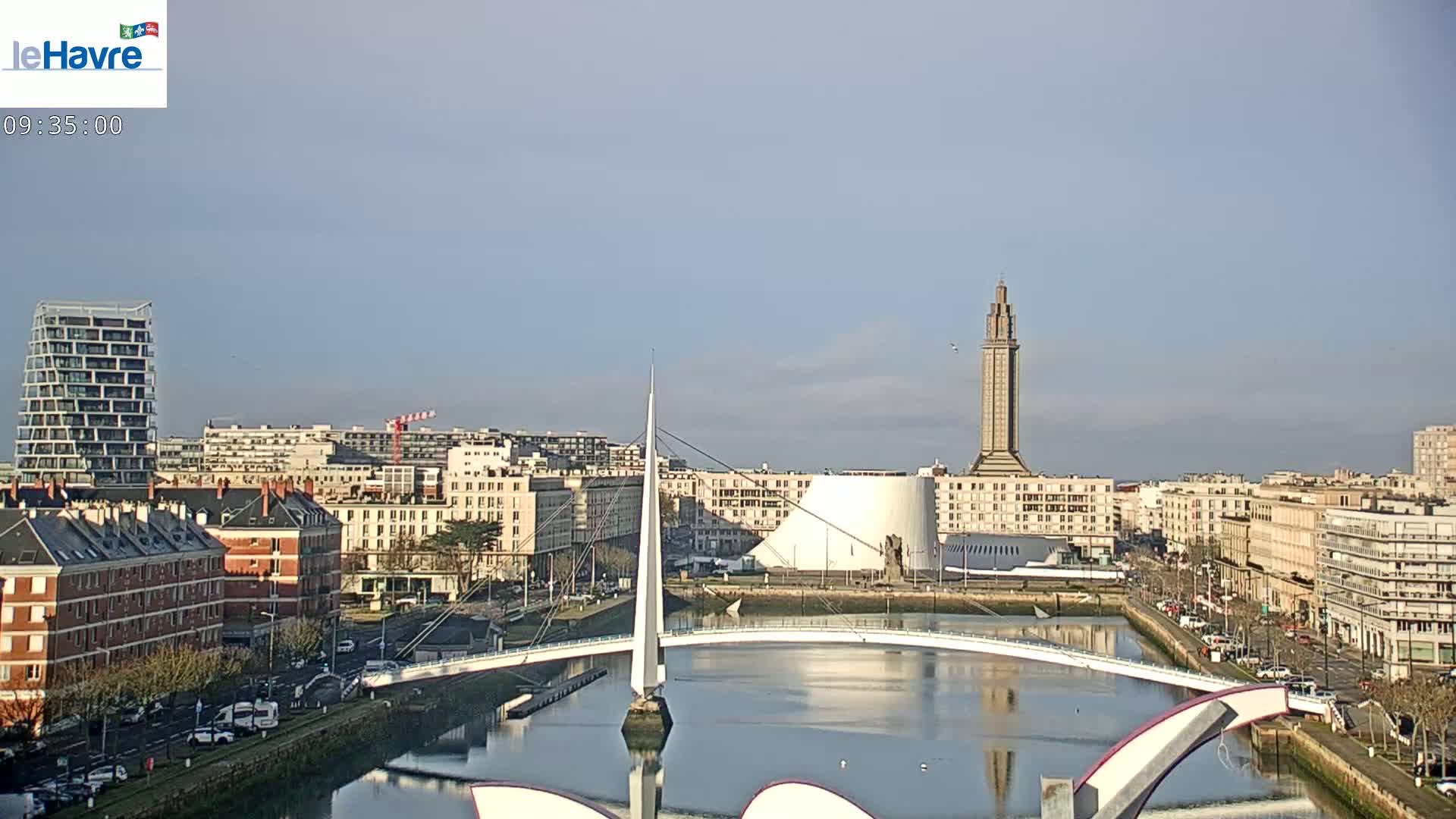 A cityscape with a modern white bridge over a calm body of water, under a clear, sunny sky, features a tall tower and various buildings in the background.