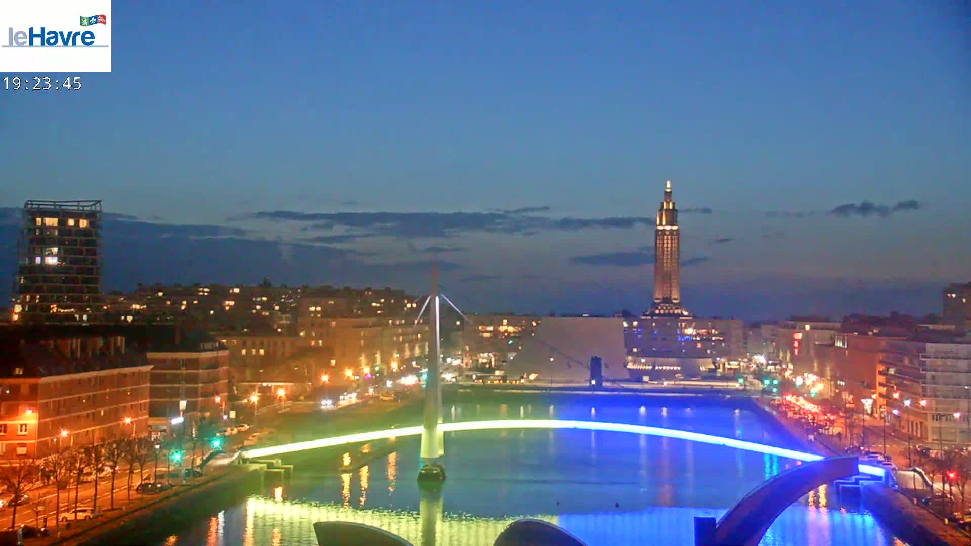 A city at night, illuminated with lights, features a bridge with blue lighting spanning a canal, with a tall building visible in the background under a twilight sky.