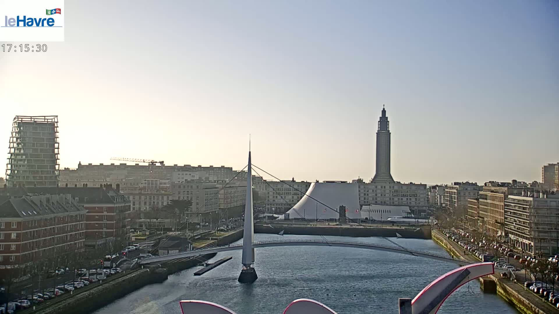 A city skyline on a clear, sunny day is shown, featuring a tall tower, modern buildings, a bridge crossing a waterway, and parked cars lining the streets.