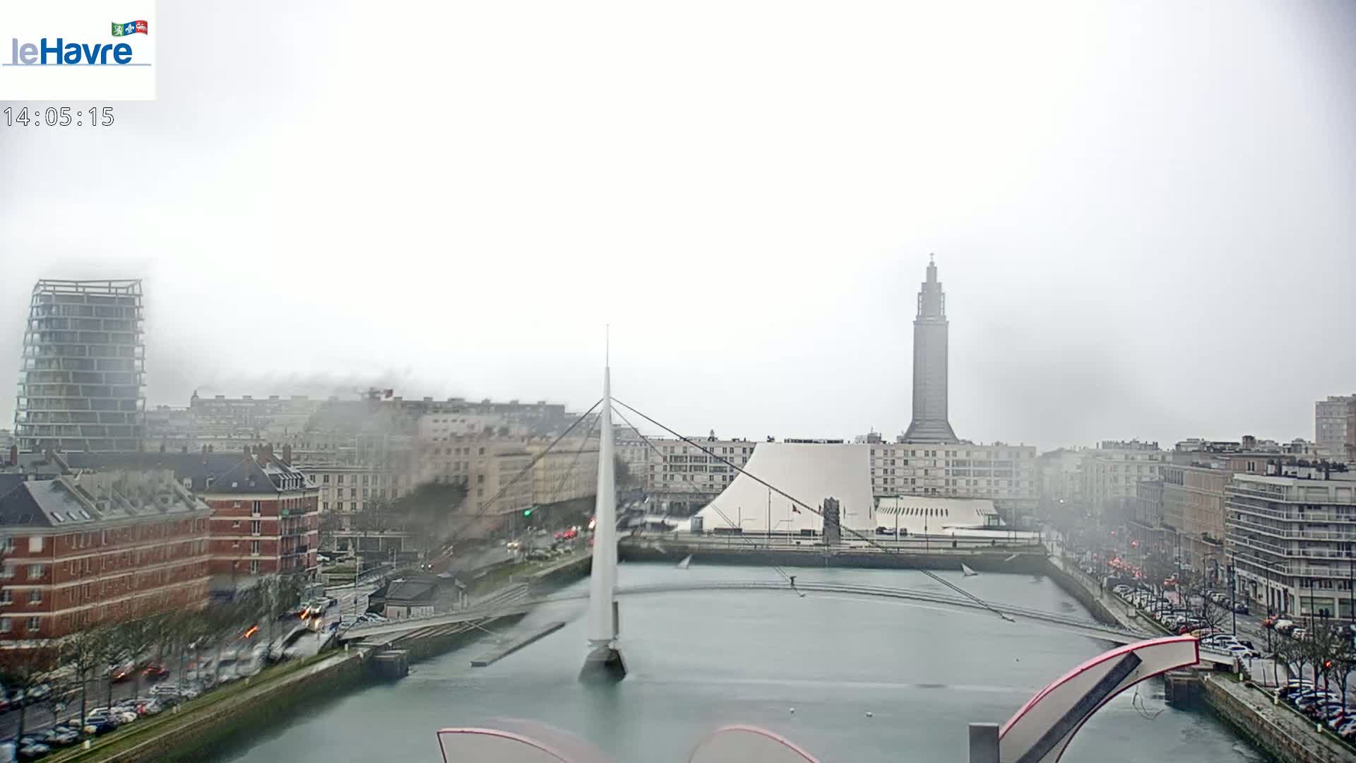 A foggy city scene shows a waterway with a bridge and several buildings, including a tall tower, under an overcast sky.