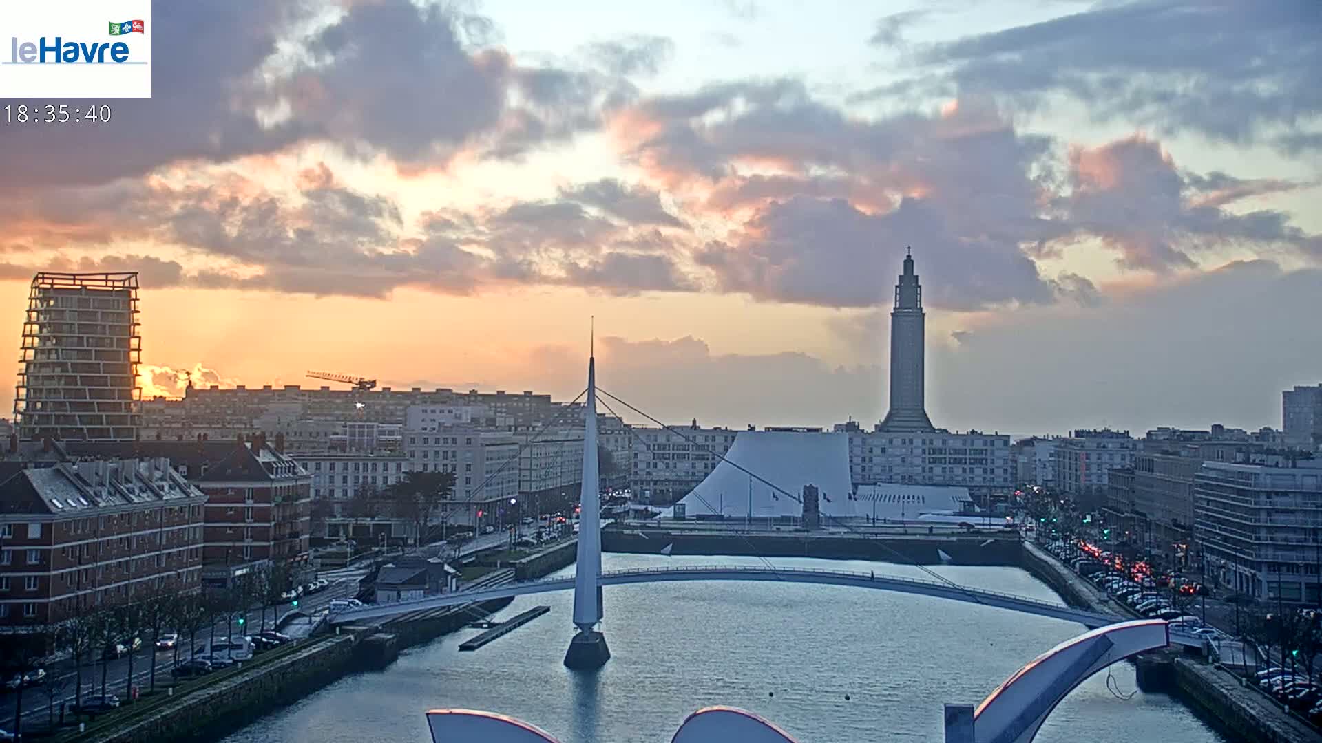 A city skyline at sunset under a partly cloudy sky, featuring a tall tower, modern buildings, a bridge over a waterway, and a sunset with orange and pink hues.