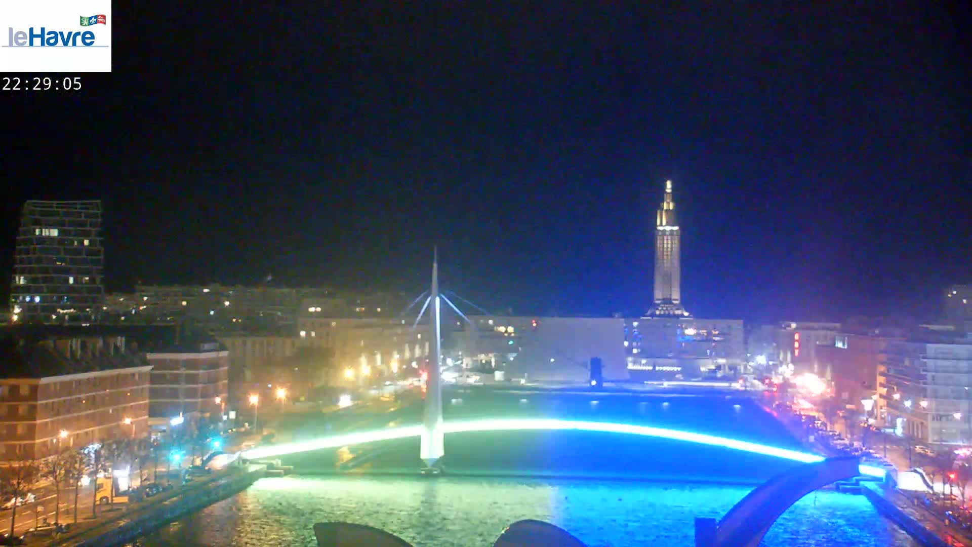 A nighttime city scene shows a bridge illuminated in blue light across a waterway, with buildings and streetlights visible along the shore under a clear, dark sky.