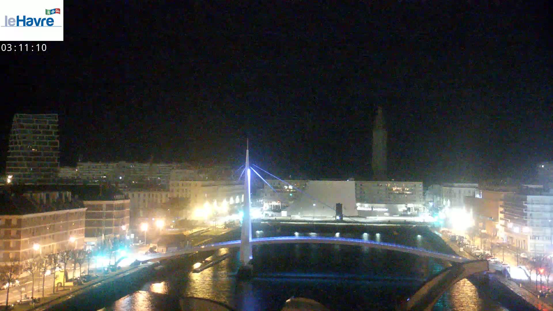 A nighttime view of a city with a modern bridge illuminated in blue light, reflecting in dark water, under a clear, dark sky.