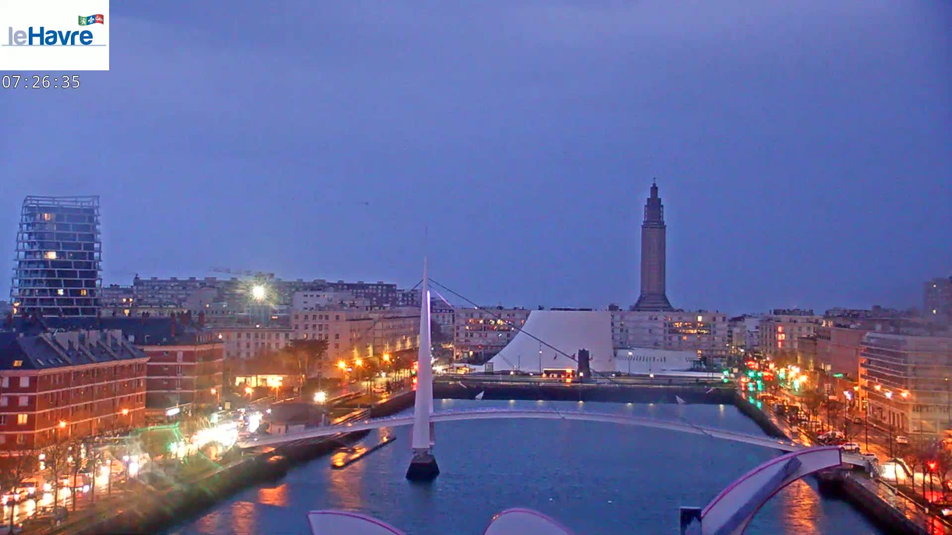 A nighttime cityscape shows a body of water with a modern white bridge, city buildings with lights, and a tall tower in the distance under a dusky sky.