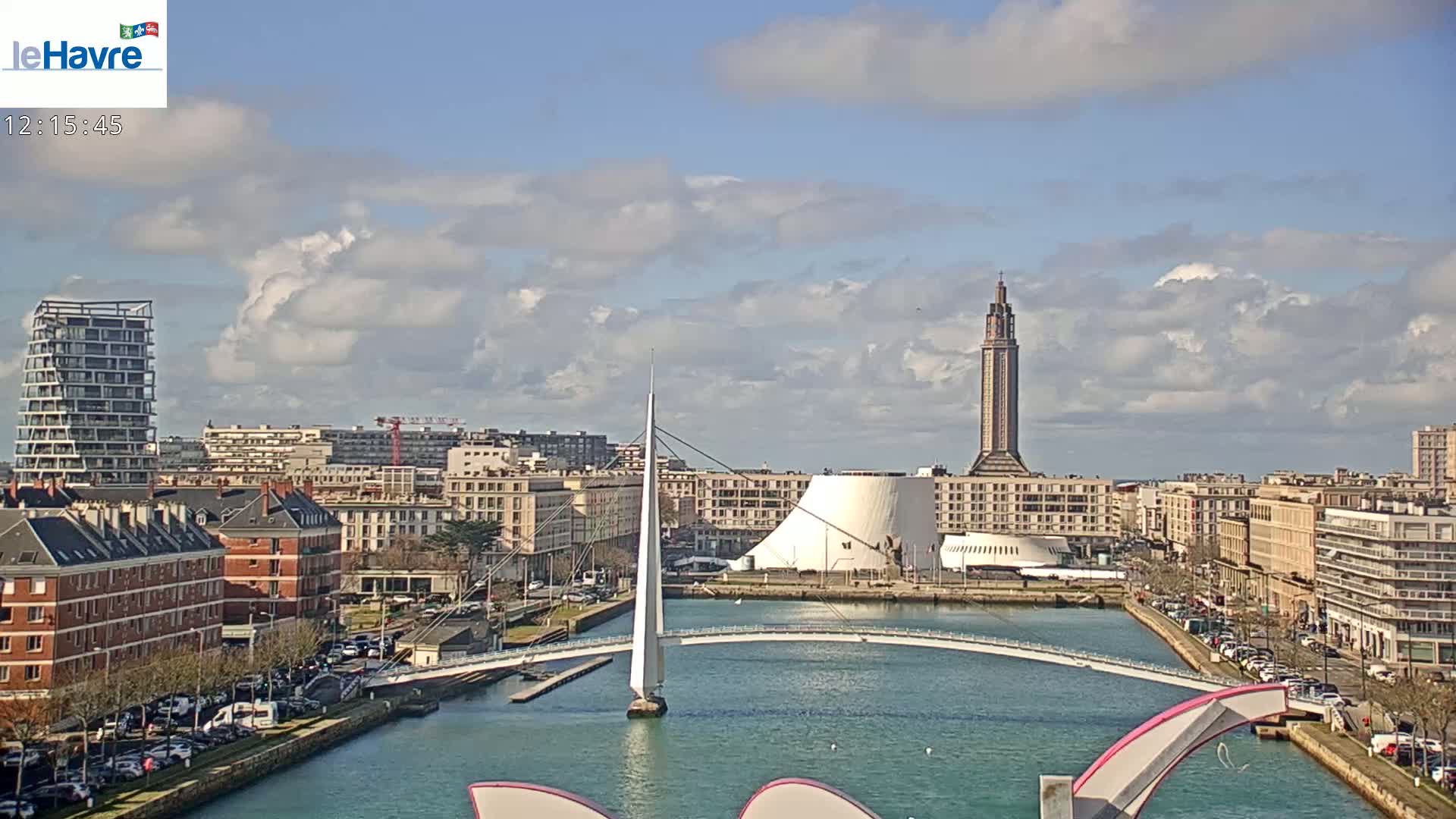 A partly cloudy day reveals a cityscape with a canal, a modern bridge, and a tall tower, all near a body of water.
