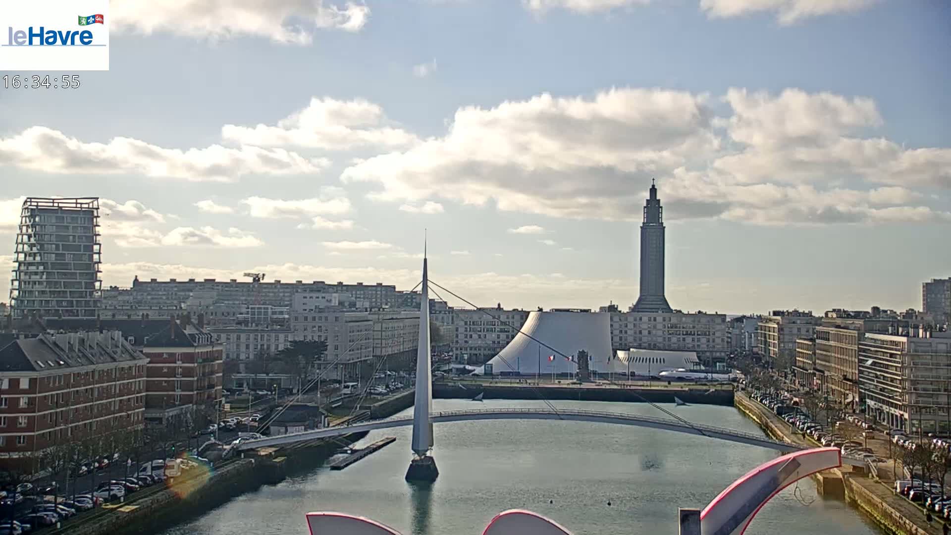A city waterfront scene under a partly cloudy sky shows a canal with a modern pedestrian bridge, buildings of varying heights, and a tall tower in the background.