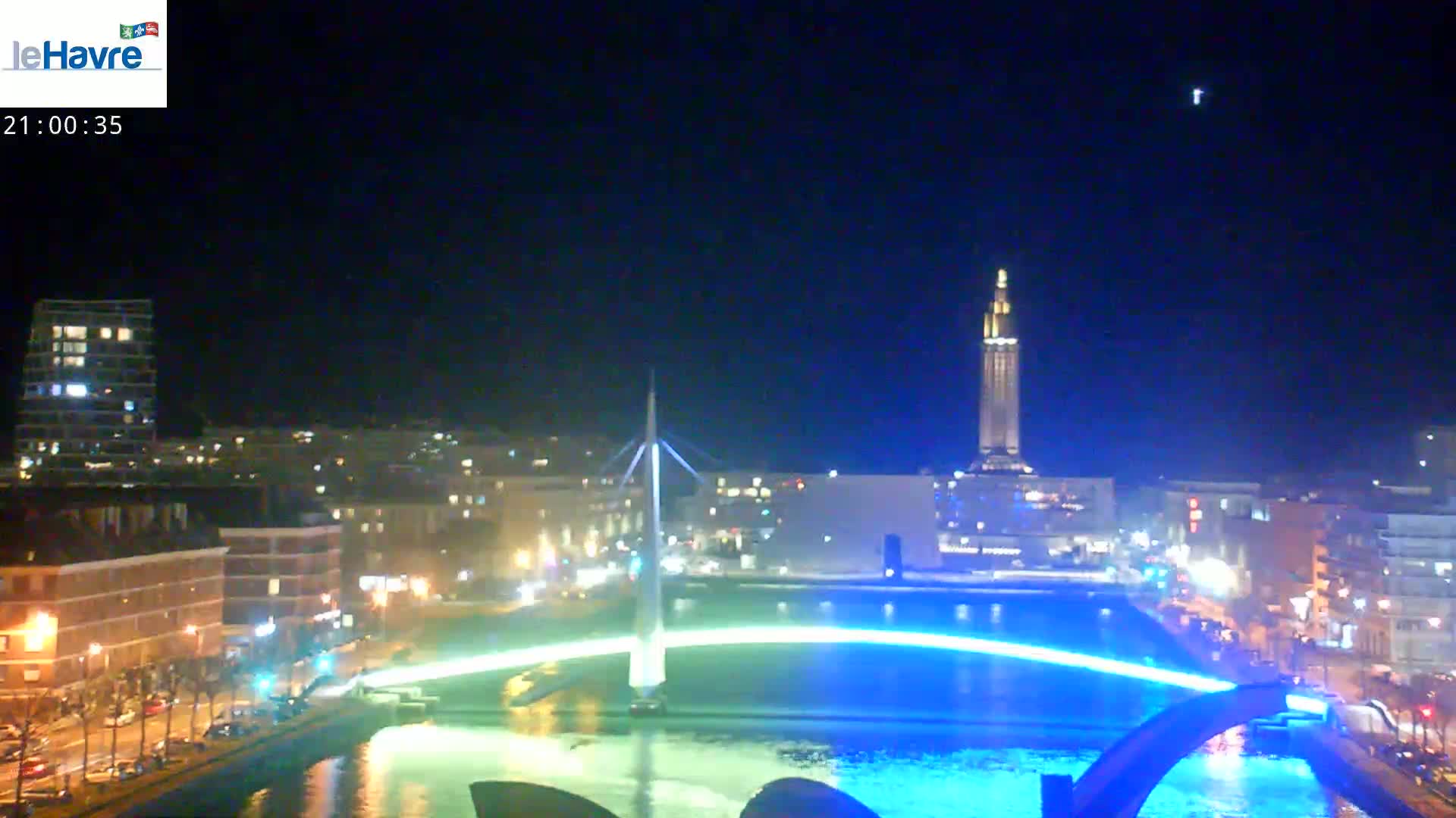 A nighttime cityscape with a bridge illuminated in blue light, reflecting in the dark water below, and a tall tower in the background under a clear, dark sky.