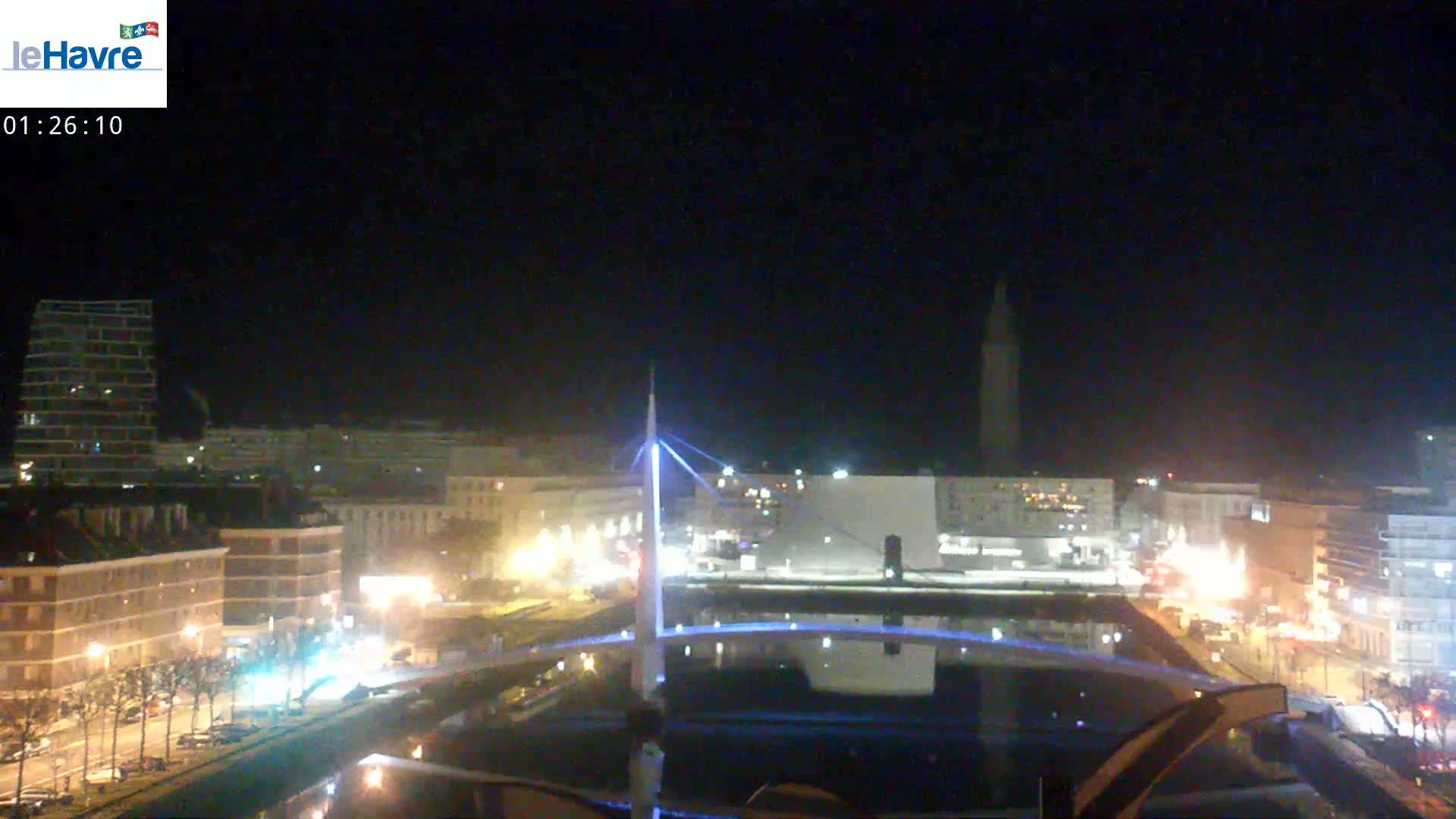 A nighttime view of a city harbor with a lit-up bridge, buildings, and streetlights, under a clear, dark sky.