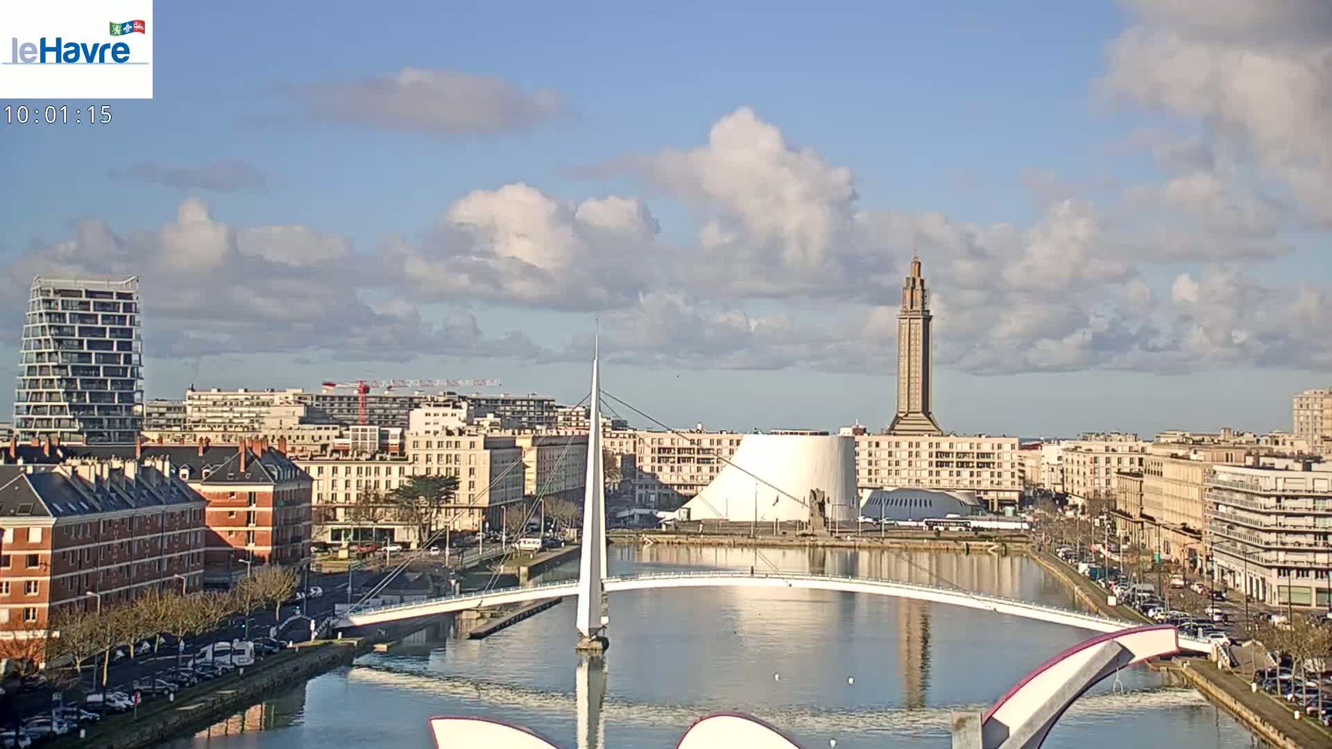 A city waterfront scene under partly cloudy skies shows a canal with a white pedestrian bridge and various buildings, including a tall tower, in the background.