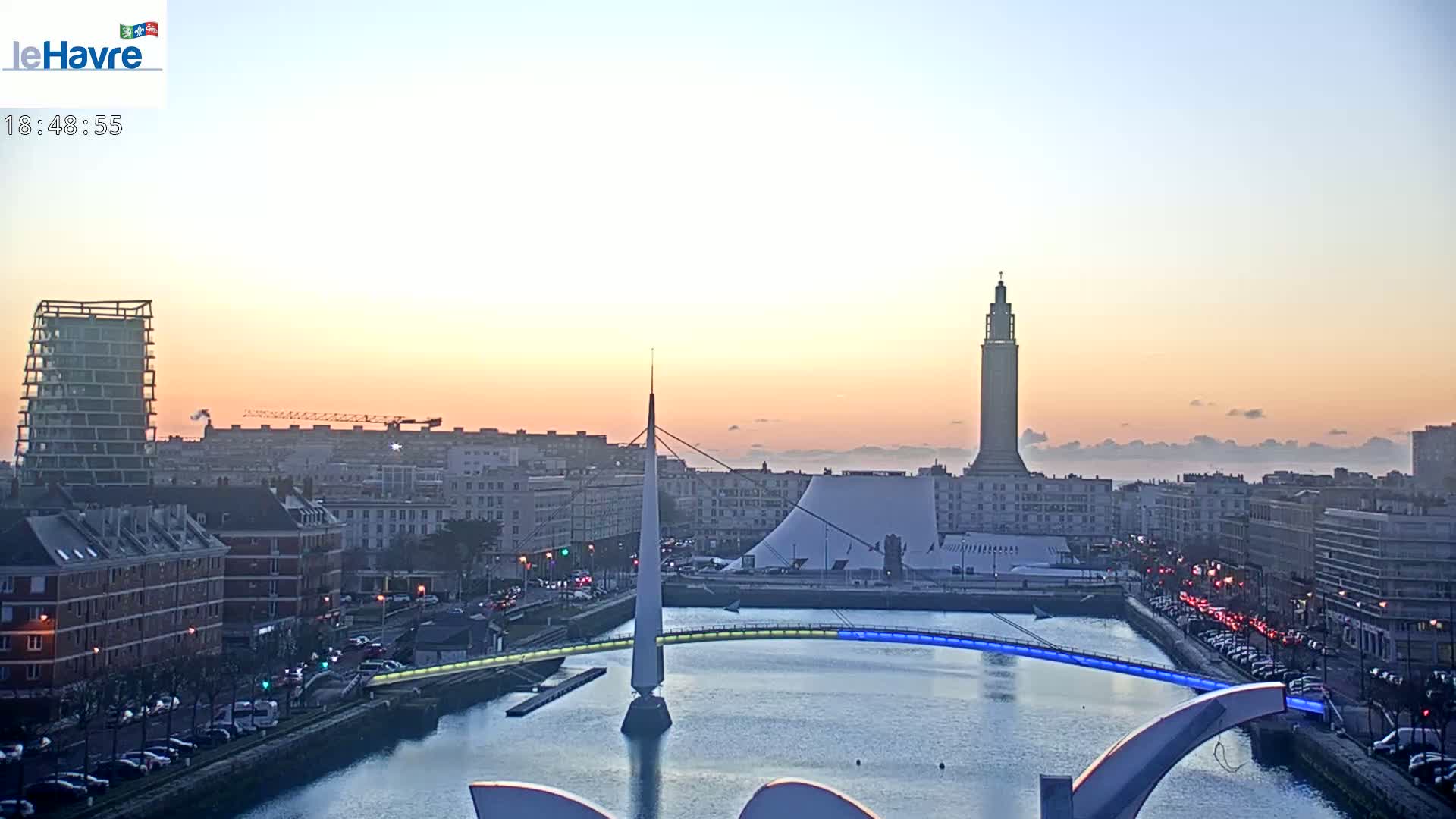 A city skyline at sunrise with a calm body of water reflecting the pale pink and orange sky, featuring a modern bridge and a tall tower.