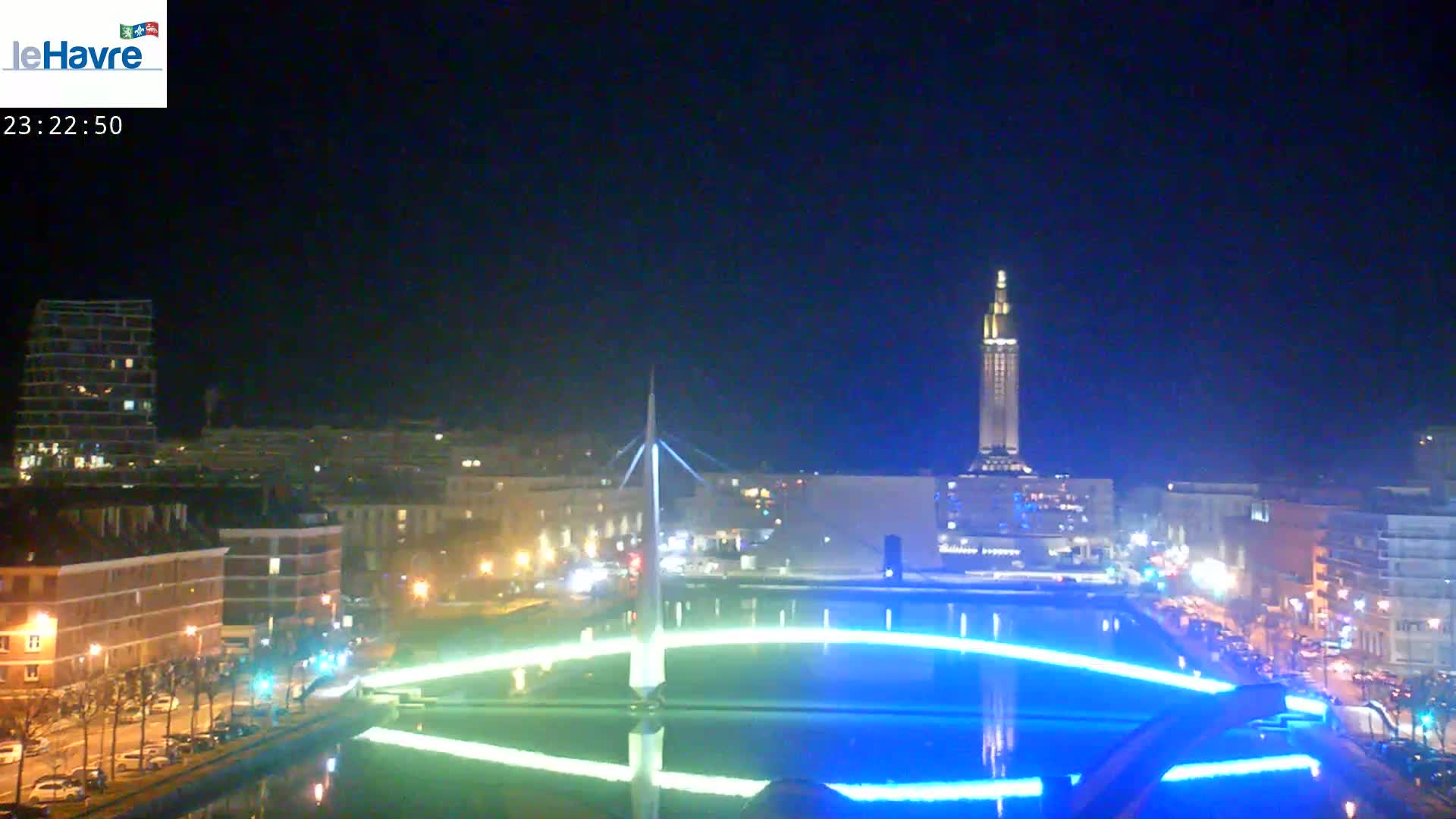 A nighttime view of a city waterfront with a tall tower in the background, illuminated by artificial lights, reflecting in a body of water with blue and green light displays.