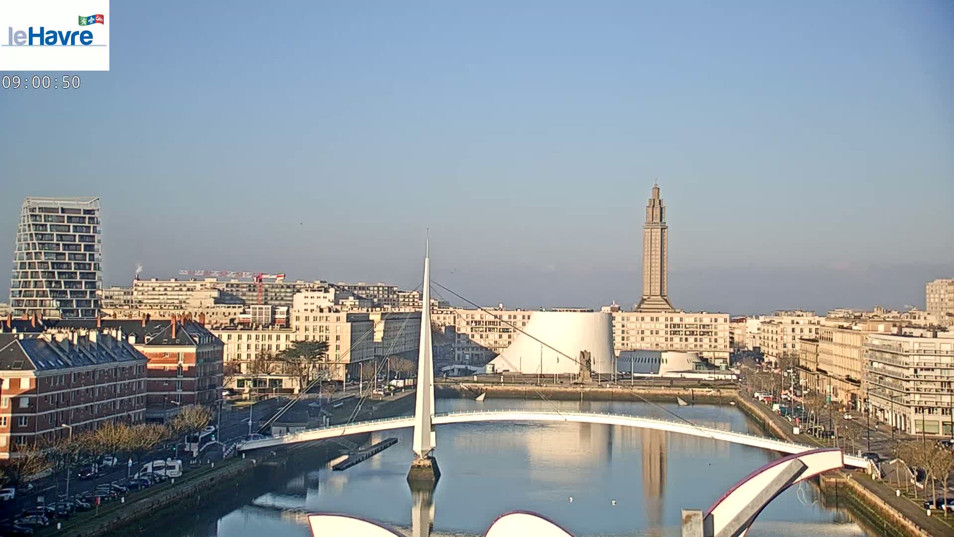 A cityscape with a modern white bridge spanning a calm body of water under a clear, sunny sky.