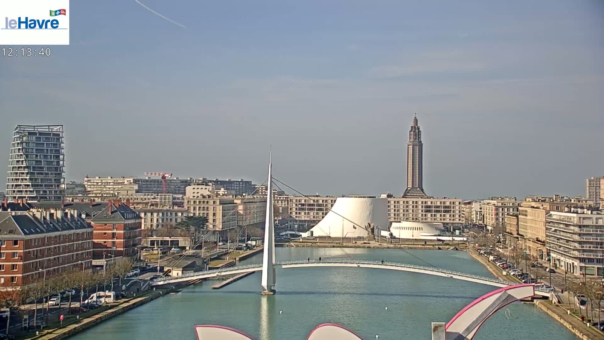 A city waterfront scene on a clear, sunny day shows a modern pedestrian bridge spanning a canal, with buildings and a tall tower in the background.