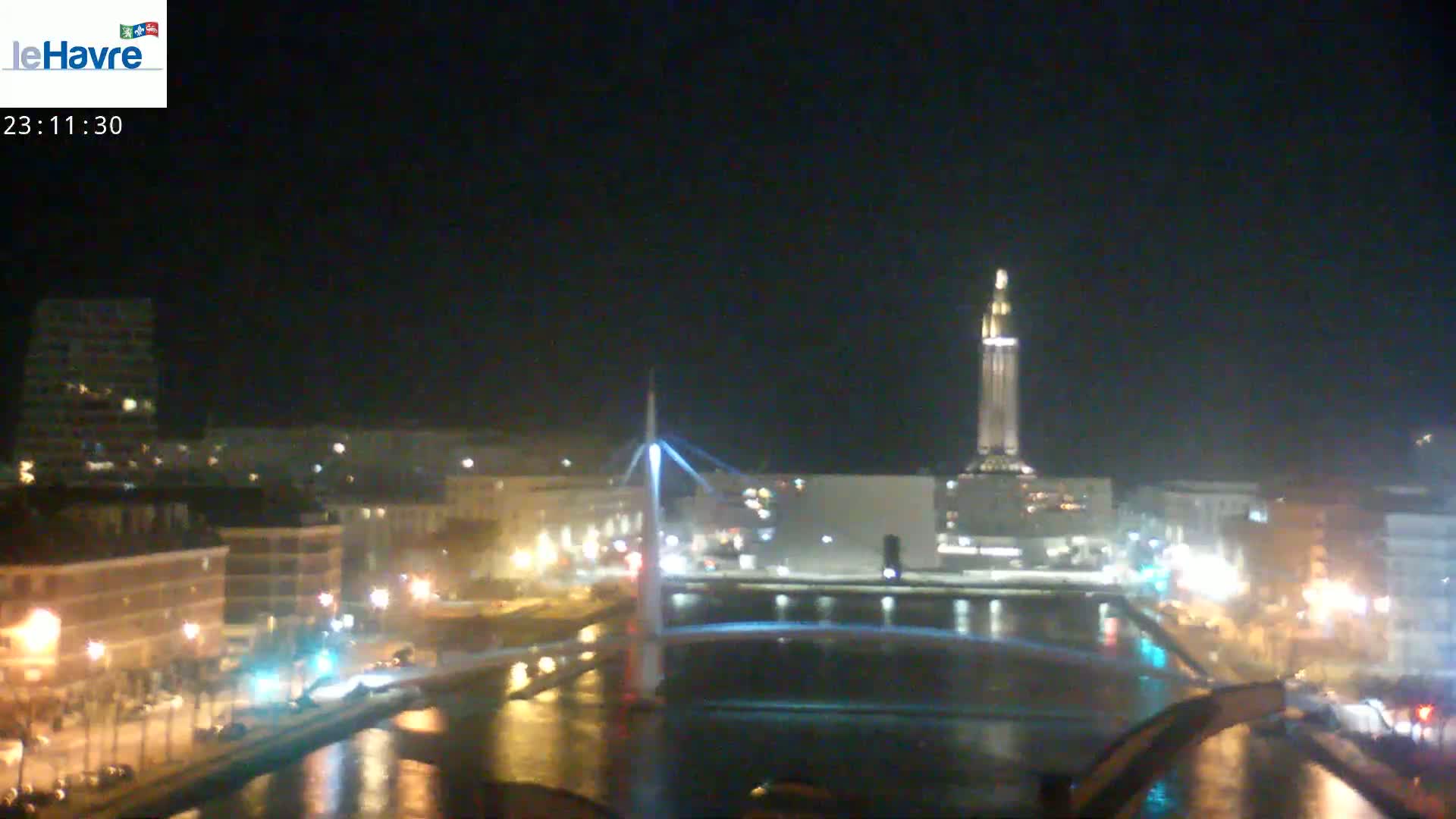 A nighttime view of a city with a bridge and tall tower over a calm body of water, under a clear sky.