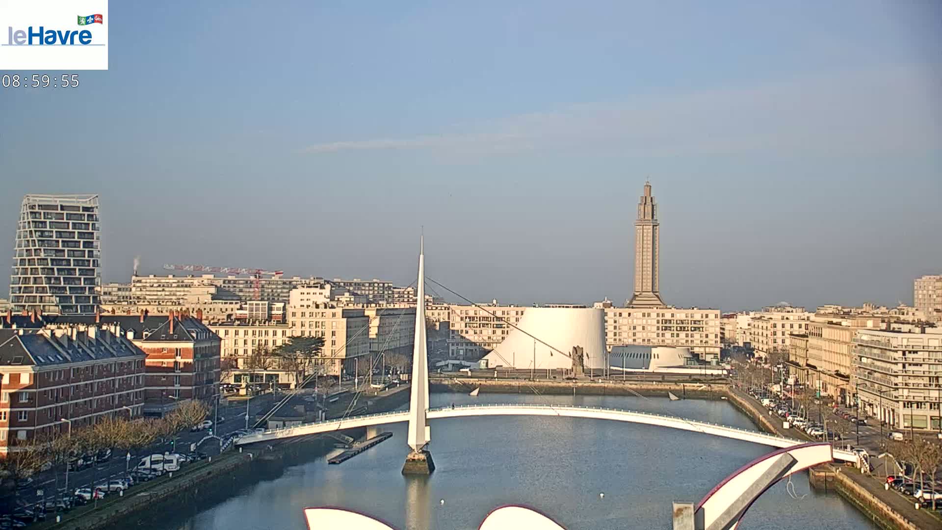 A city waterfront on a sunny day shows a modern bridge spanning a calm waterway, with buildings, including a tall tower, lining the shore.