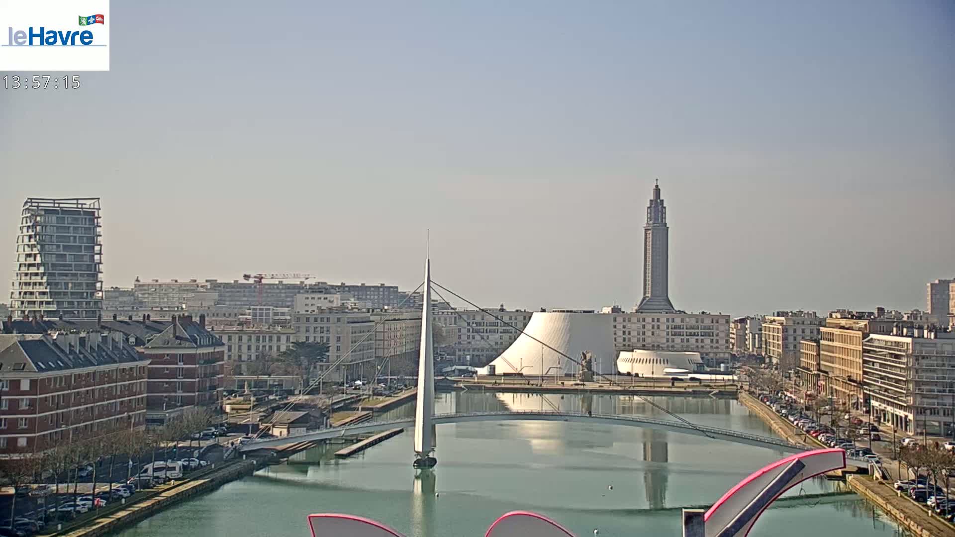 A city harbor scene on a sunny day shows a canal with a modern footbridge, buildings of various architectural styles, and a tall tower in the distance.