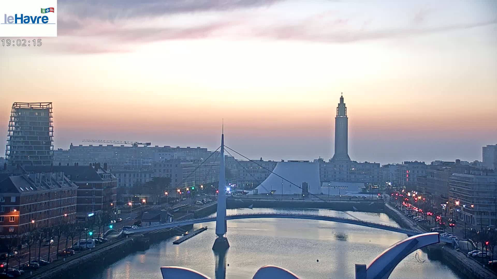 A city skyline at sunset, featuring a slender bridge over a calm waterway, a tall tower, and modern buildings, under a hazy, pastel sky.