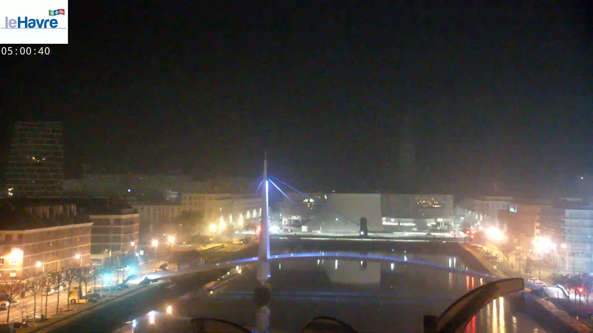 A nighttime view of a city with a lit-up bridge spanning a canal, streetlights illuminating buildings and roads along the waterfront under a dark, clear sky.