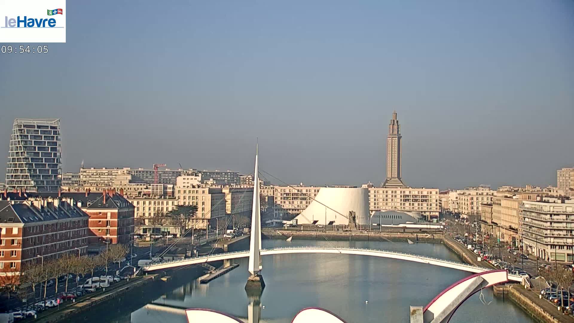 A city waterfront with a modern bridge, buildings, and a tall tower is visible under a clear, sunny sky.
