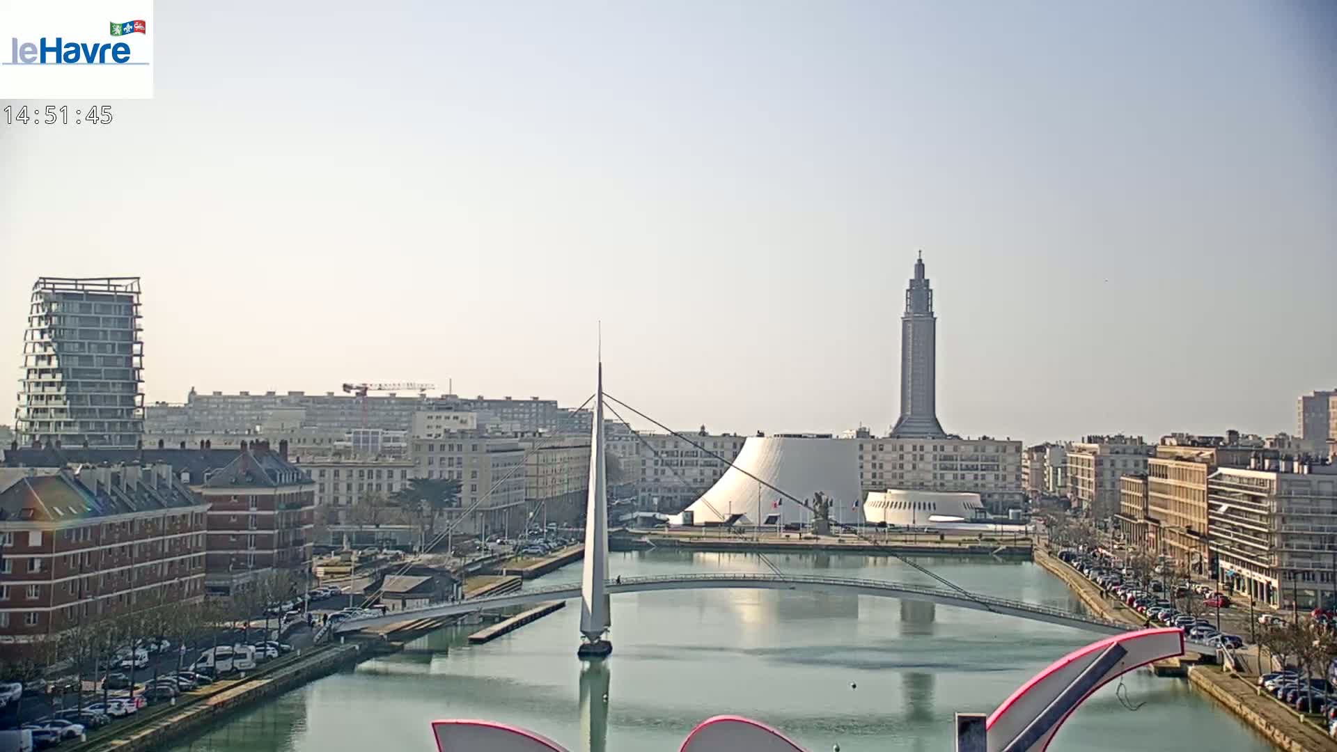 A city waterfront on a sunny day shows a canal with a modern pedestrian bridge, buildings lining the canal, and a tall tower in the background.