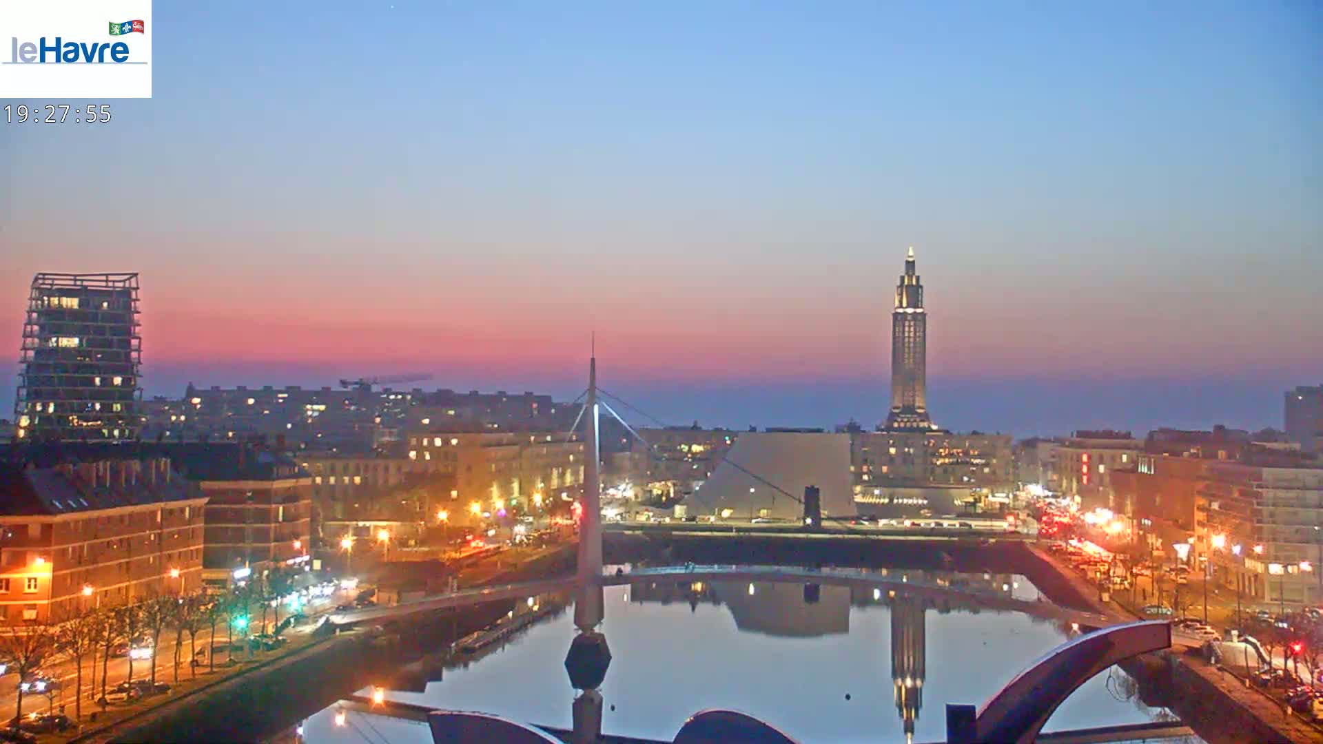 A city skyline at twilight, with a calm body of water reflecting the lights of buildings and a bridge, under a pinkish-purple sky.