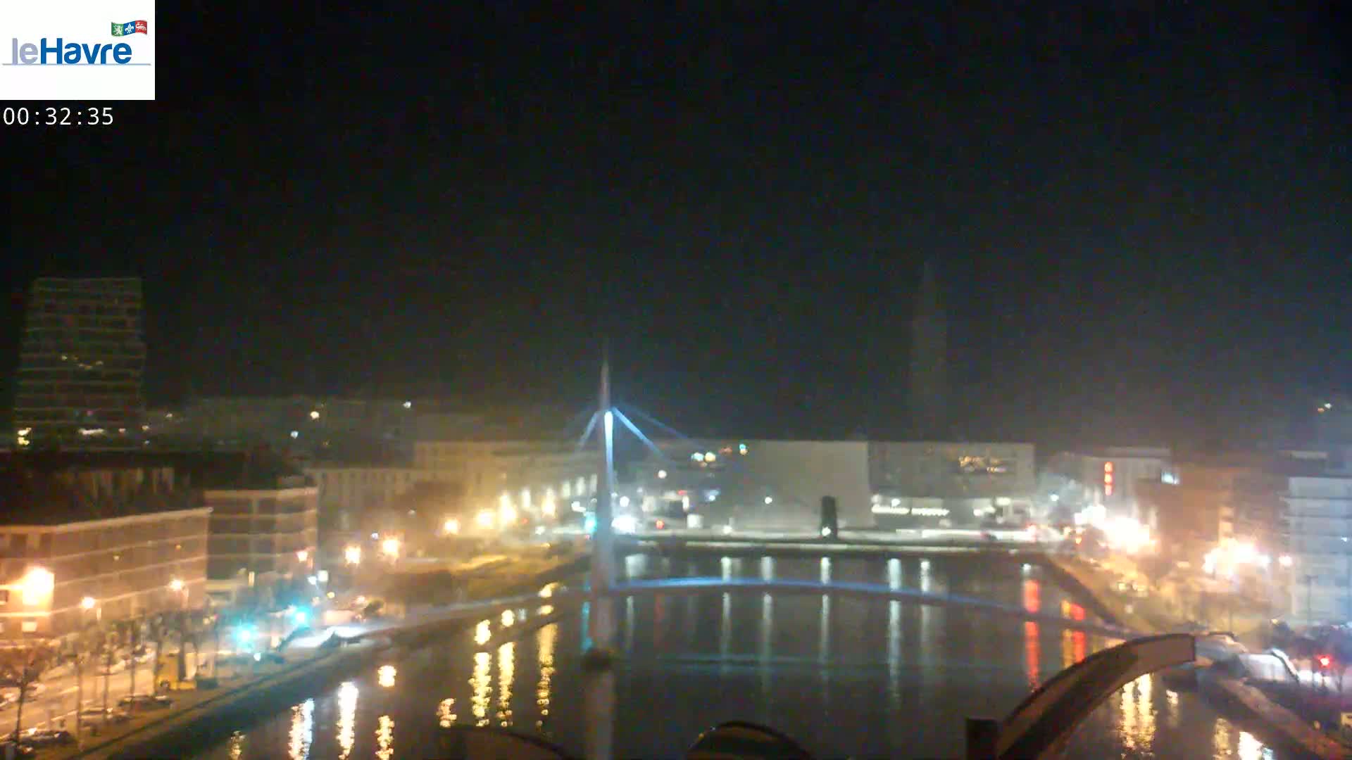 A nighttime view of a city with a lit-up bridge spanning a calm river, under a dark, clear sky.