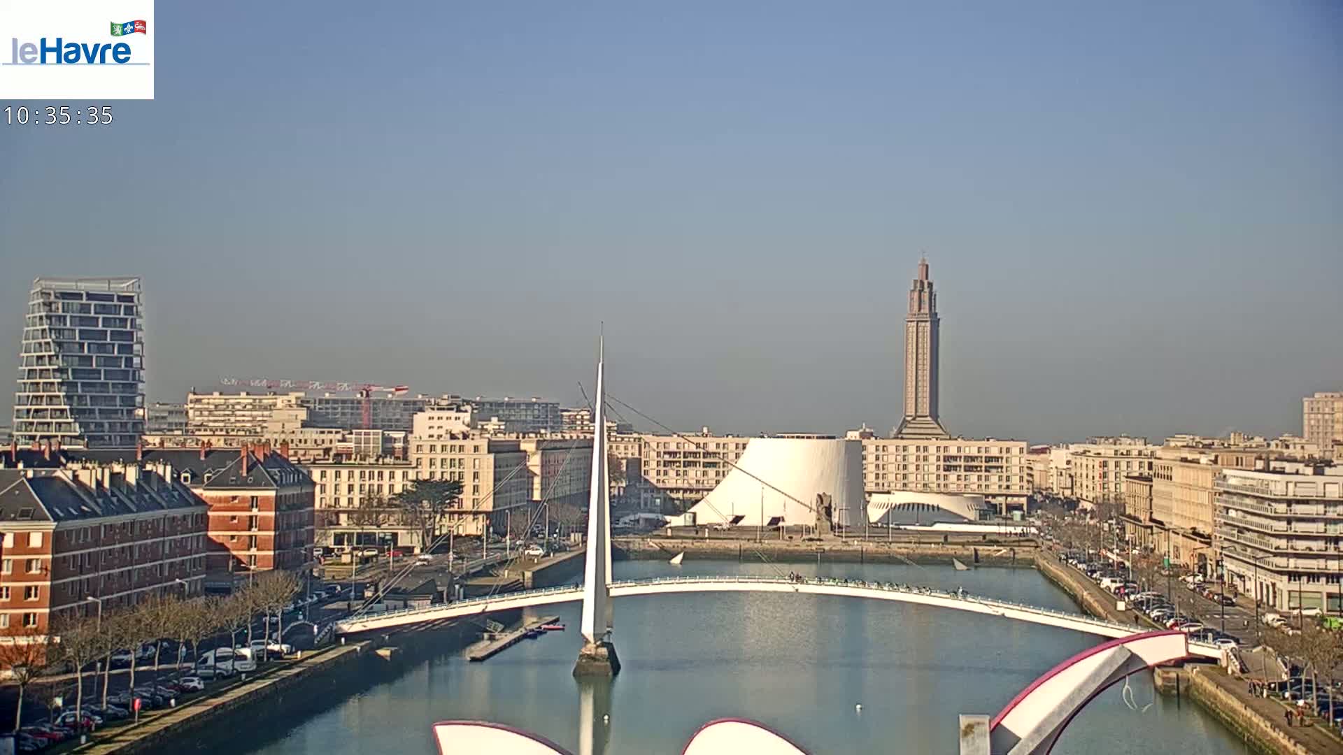 A sunny cityscape with a modern white bridge over a waterway, featuring several buildings, including a tall tower, under a clear sky.