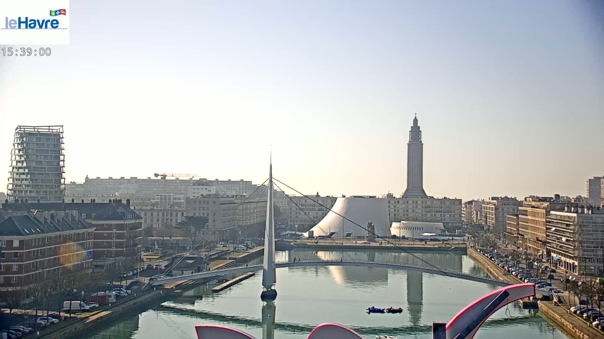 A sunny cityscape shows a waterway with a sailboat and bridge, buildings lining the waterfront, and a tall tower in the background.