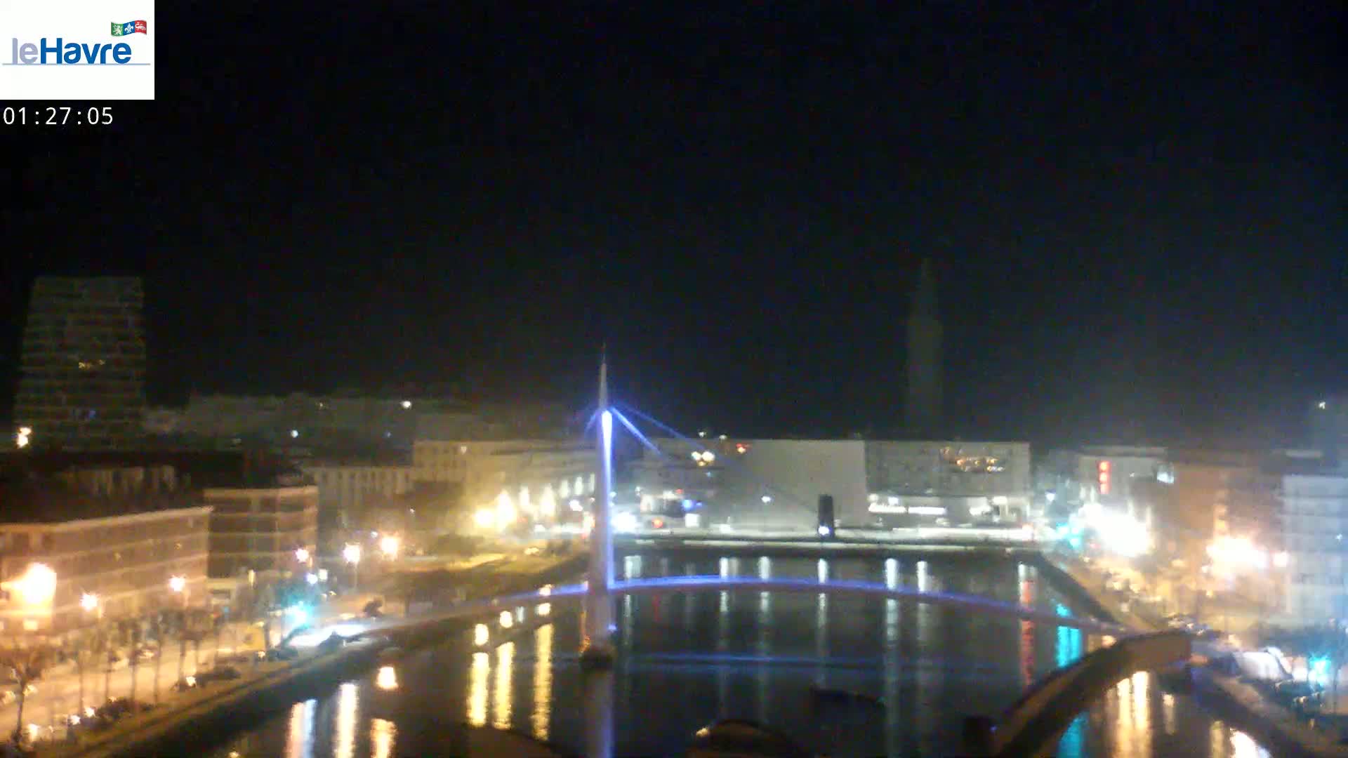 A nighttime view of a city with a bridge illuminated in blue light, reflecting on calm water.